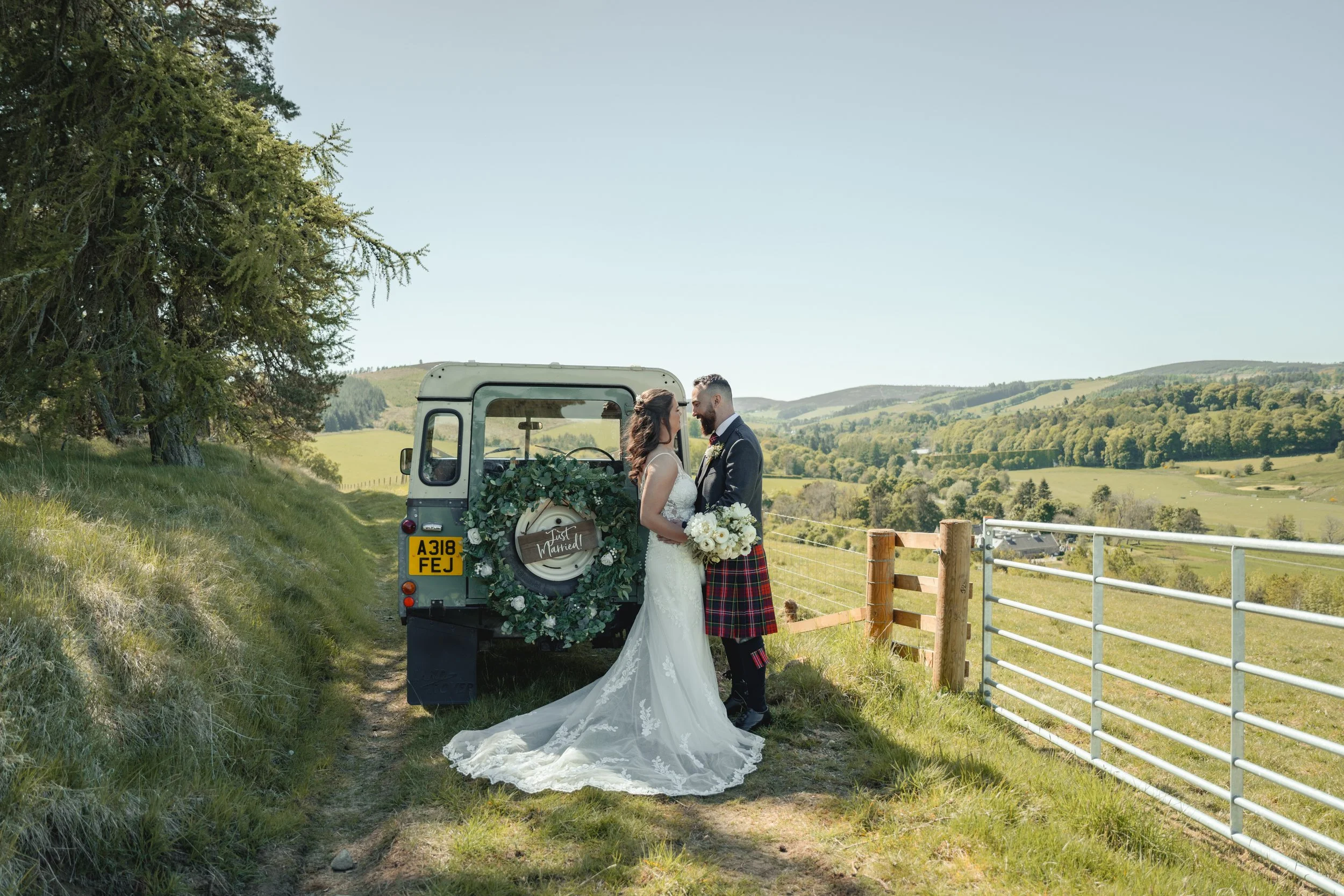 A bride and groom in wedding attire standing close together outdoors next to a vintage trailer decorated with greenery, flowers, and a 'Just Married!' sign. They are in a rural setting with rolling hills and trees in the background.
