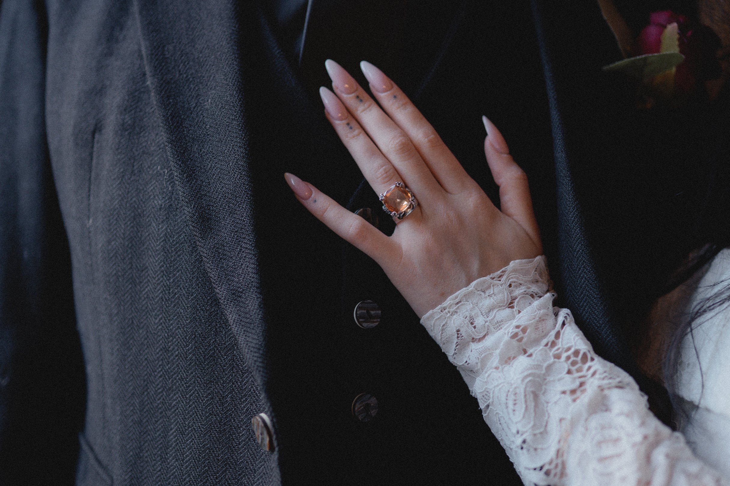 Close-up of a woman's hand with a large gemstone ring resting on a man's suit, wearing a lace sleeve
