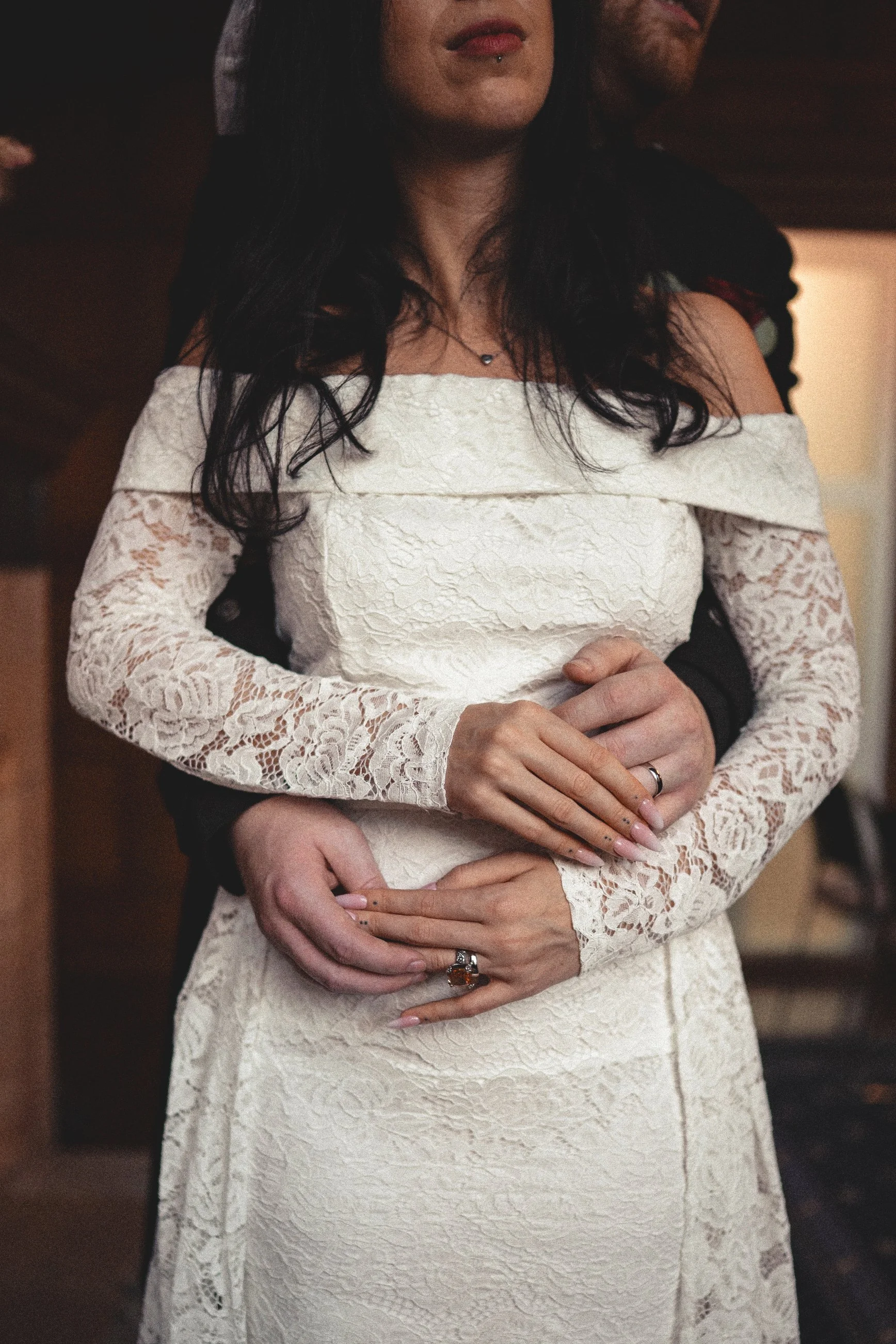 Close-up of a woman in a white lace off-shoulder dress and a man hugging her from behind.
