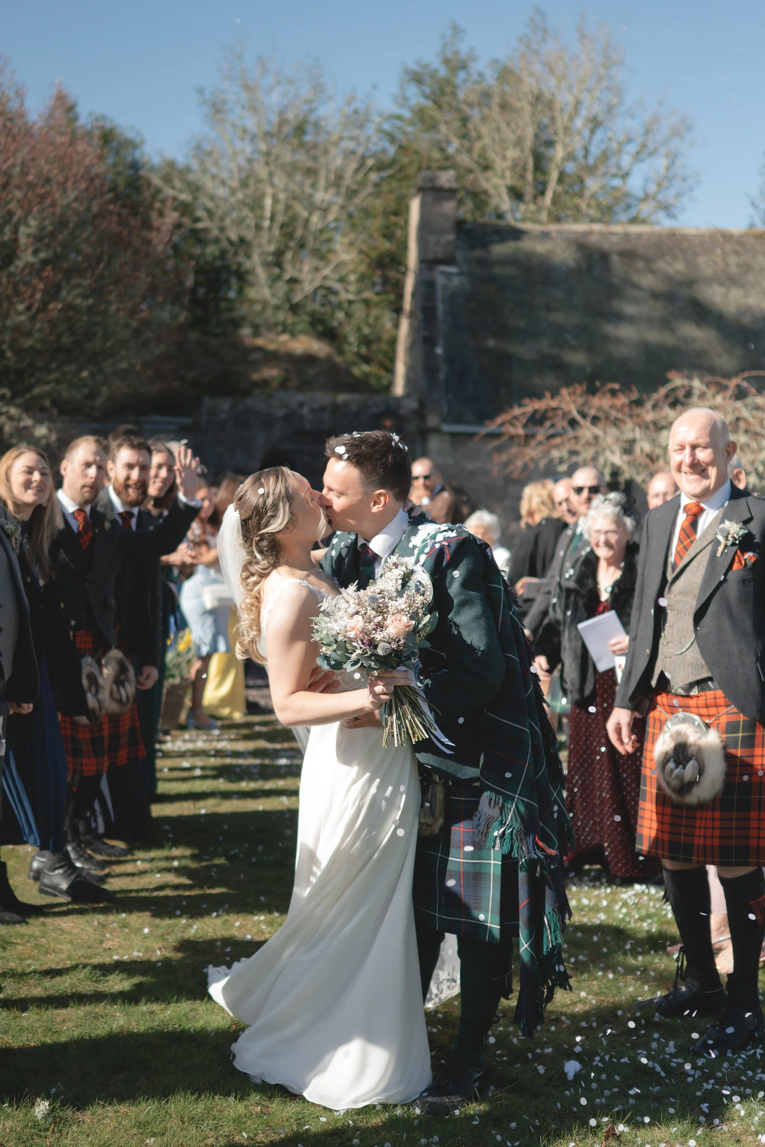 A bride and groom kiss during their outdoor wedding ceremony, surrounded by friends and family, with some guests wearing traditional Scottish attire.