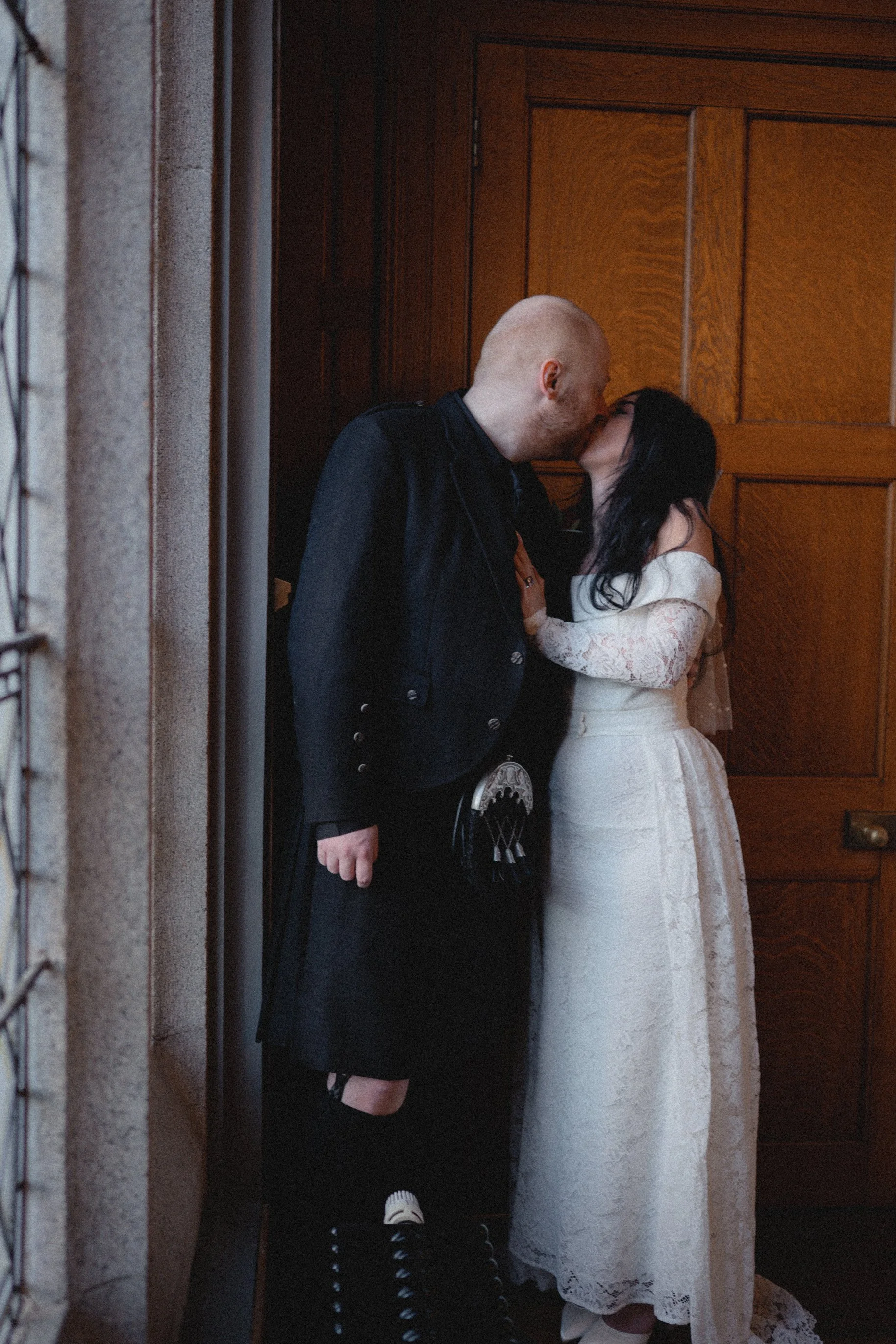 A couple sharing a kiss indoors, with the man dressed in a black jacket and kilt, and the woman in a white lace wedding dress, standing close to a wooden door.