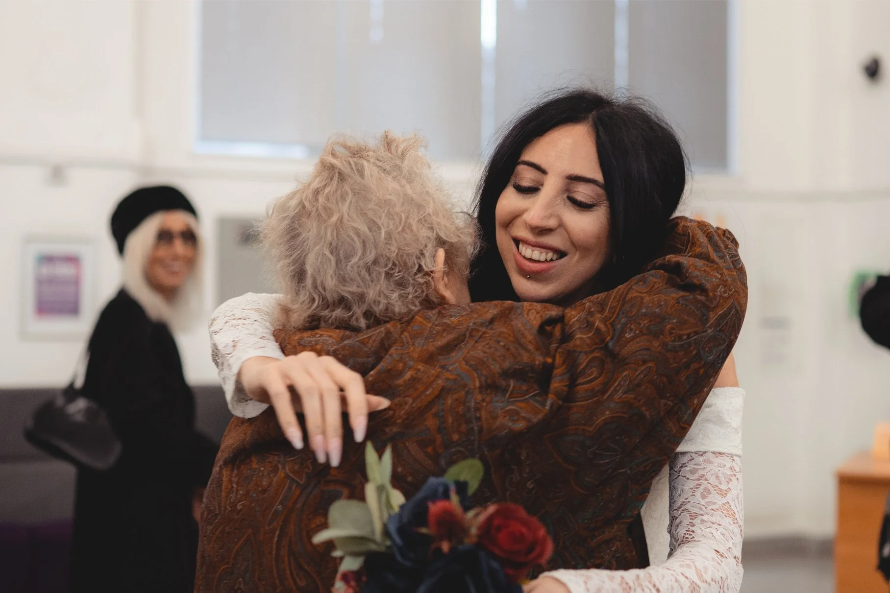 Two women are hugging each other in a joyful and emotional embrace in an indoor setting. One woman has curly blonde hair and is holding a bouquet of flowers, while the other woman has dark hair and a bright smile.