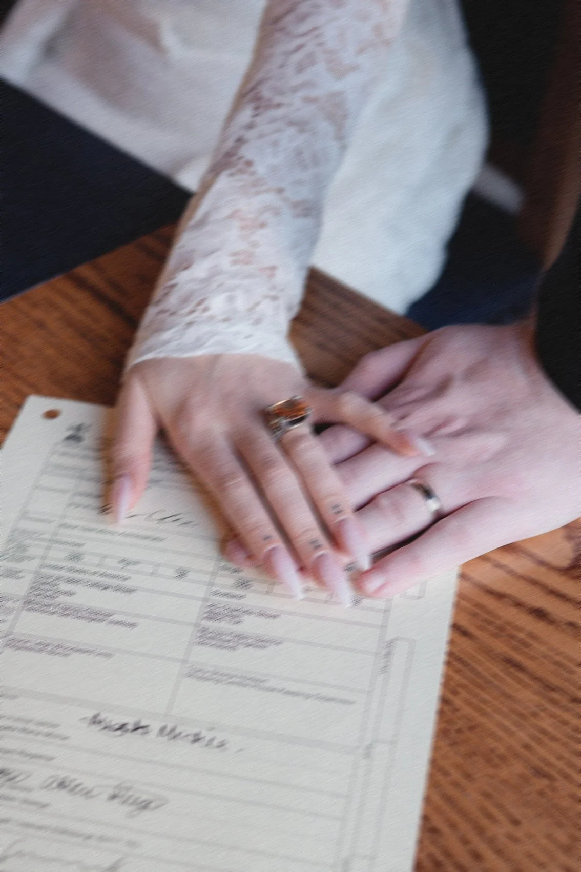 Close-up of hands, one with a wedding ring, resting on a table over a form or application, with wooden surface visible.