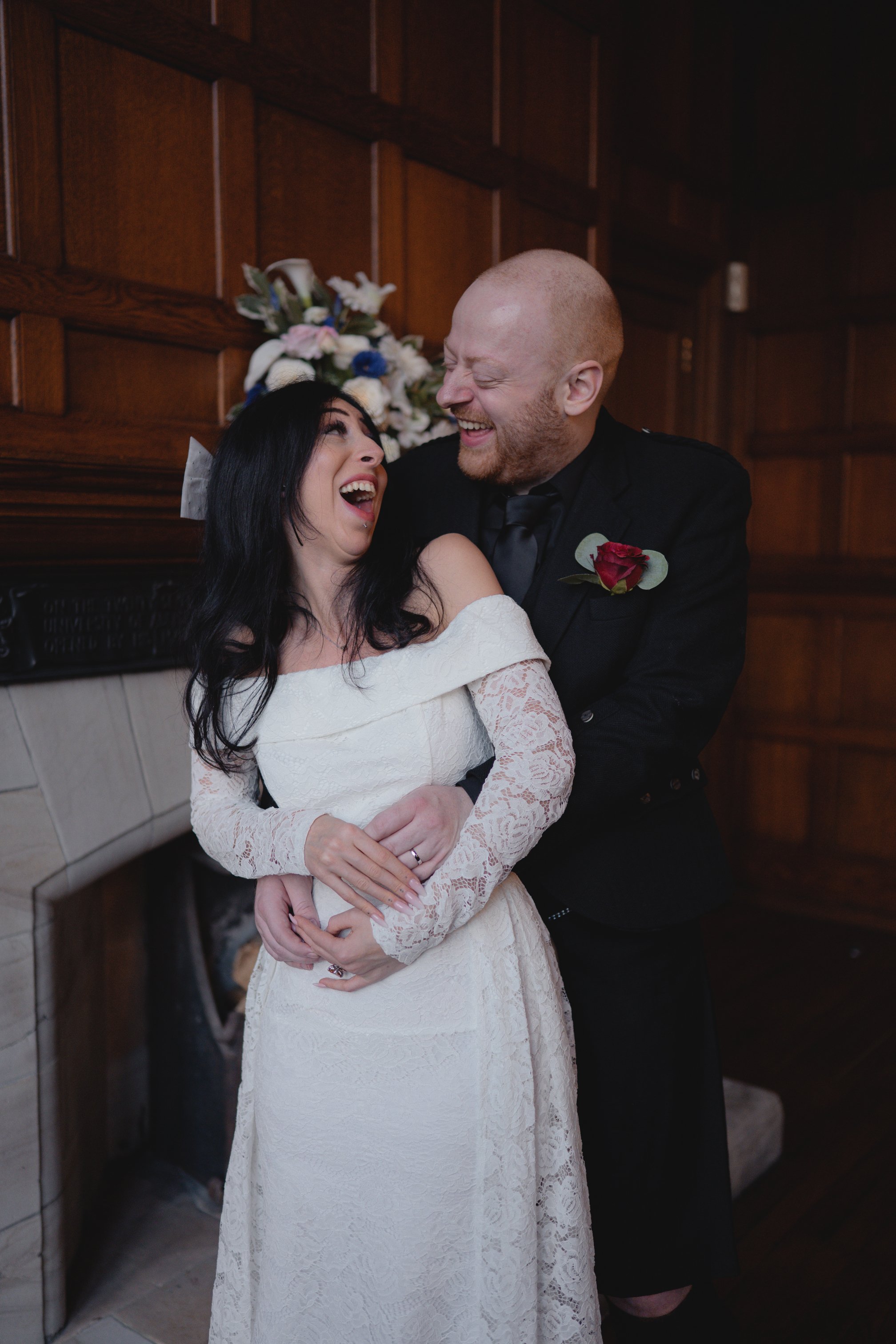 A woman in a white lace wedding dress and a man in a black suit are laughing and holding each other in front of a wooden wall with a floral arrangement behind them.