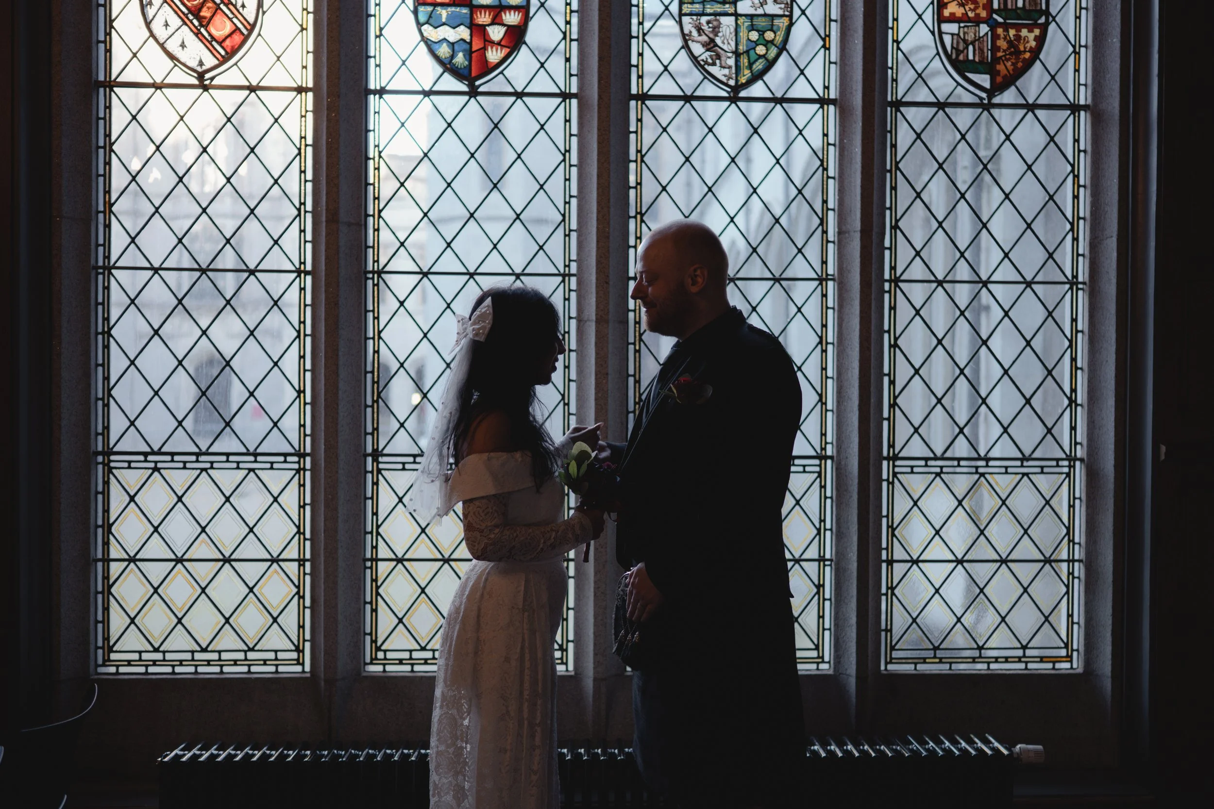 Silhouetted bride and groom standing face-to-face in front of stained glass window during wedding ceremony.