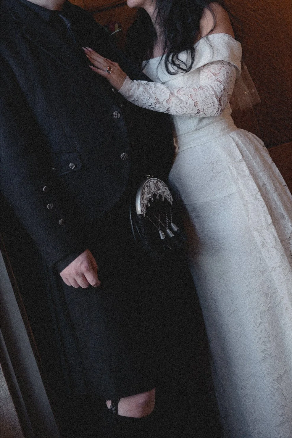 A couple dressed in wedding attire, standing close together. The woman is in a white lace wedding dress, and the man is in a black suit with a matching bow tie.