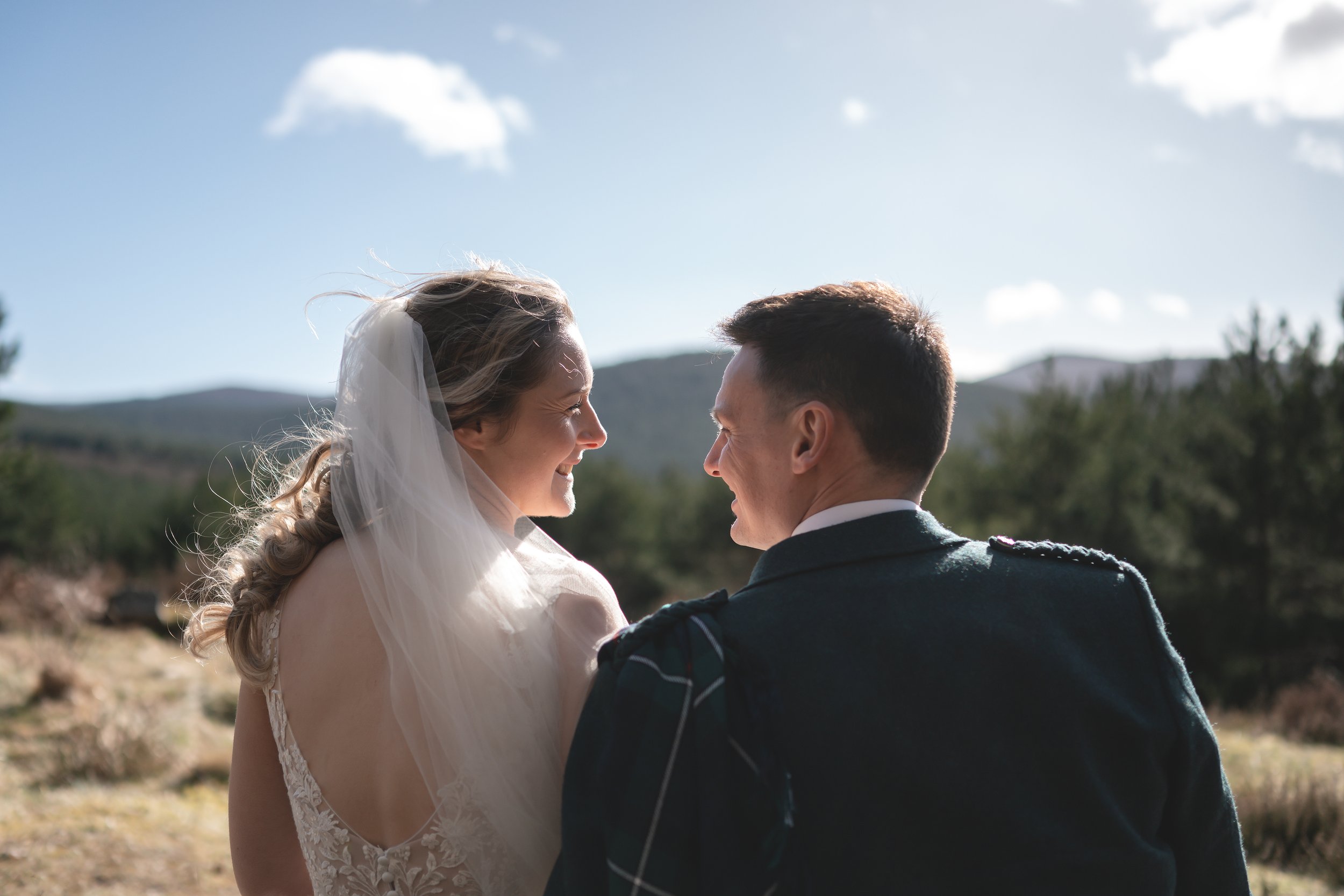 A bride and groom smiling at each other outdoors on their wedding day with mountains and trees in the background.