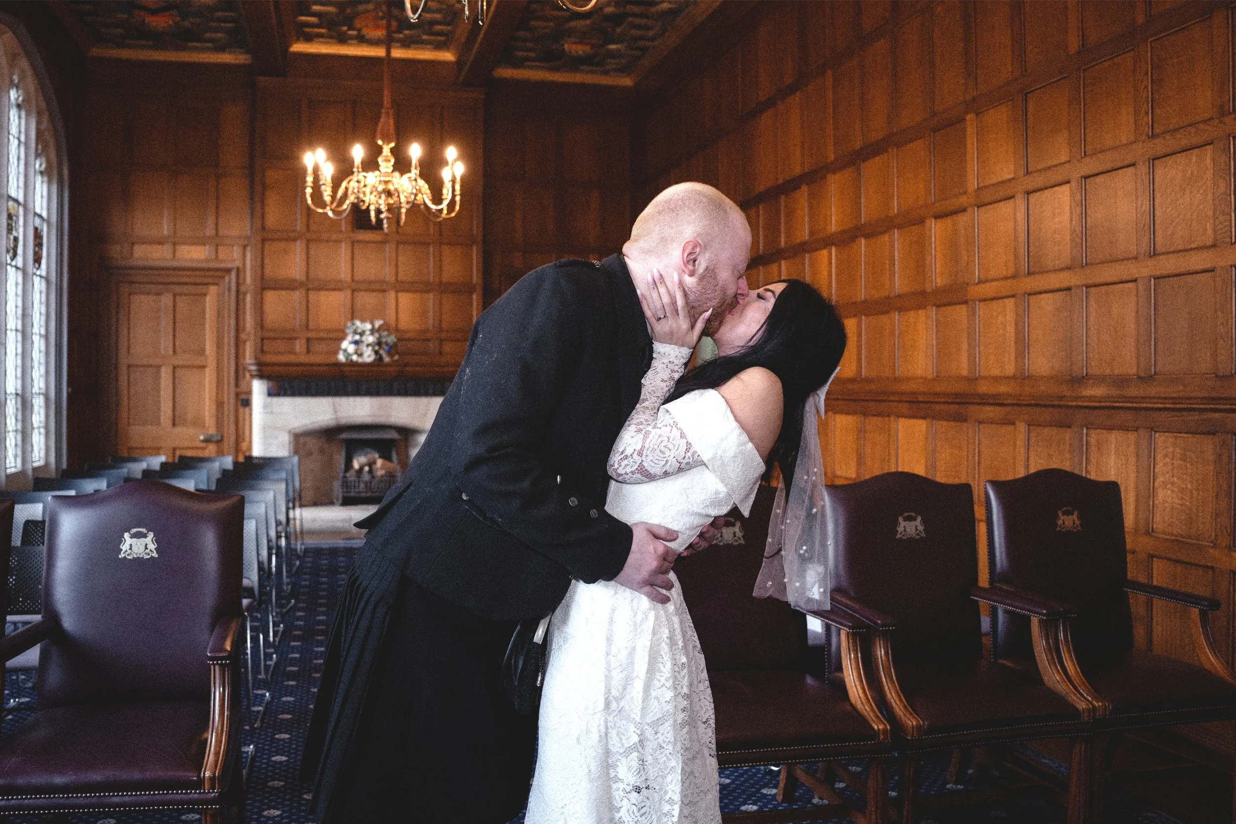 A couple kissing in an ornate wood-paneled room with high ceilings, chandelier, and chairs.