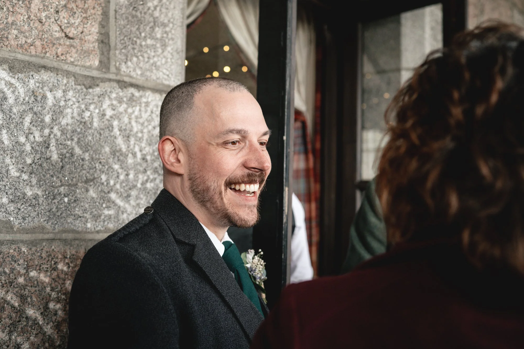 A man in a dark suit and tie is smiling and talking to a woman with curly hair at what appears to be a social event or wedding reception.