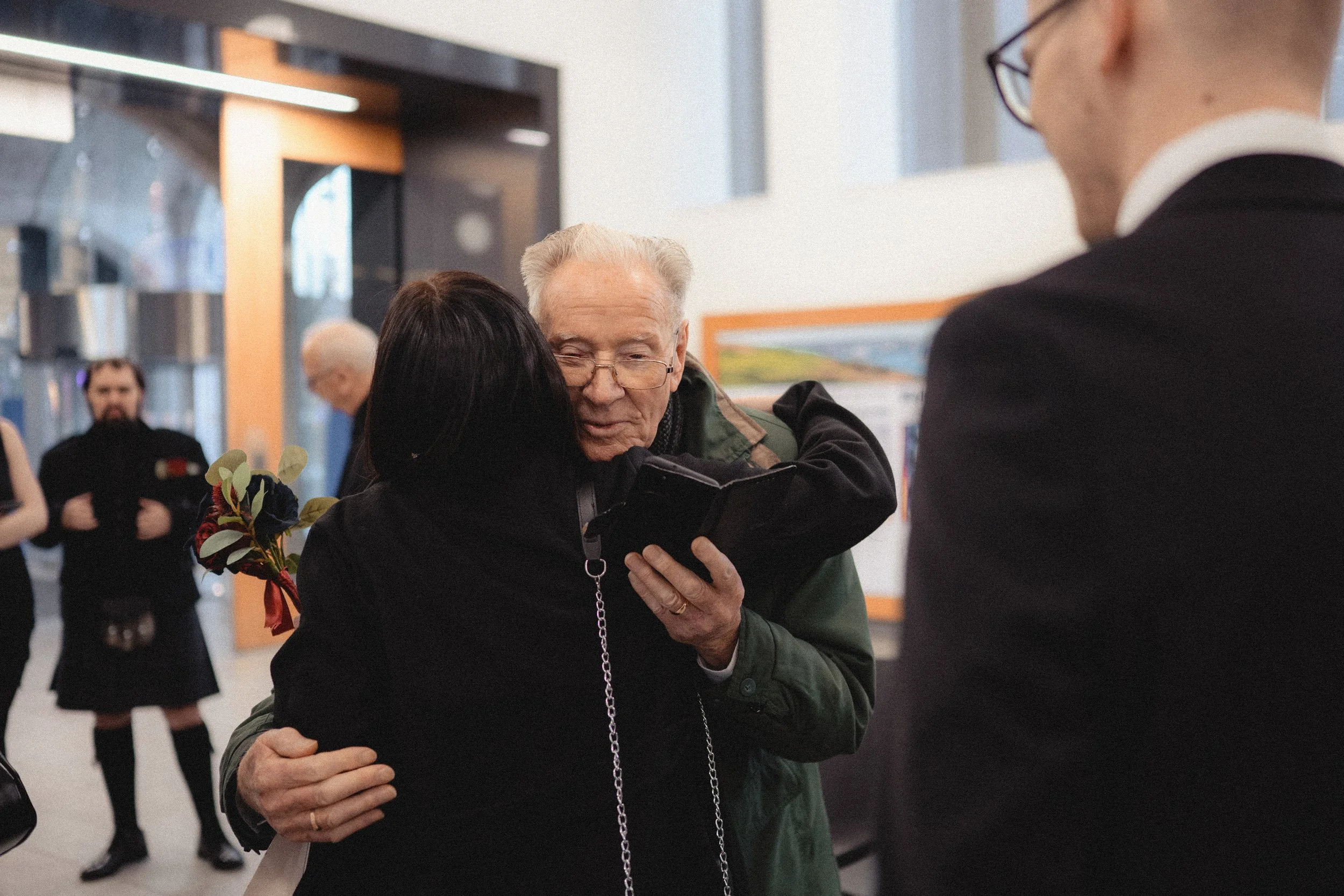 An elderly man and a woman hugging while looking at a smartphone, with a man in a suit observing them, in a public indoor space with people and artwork in the background.
