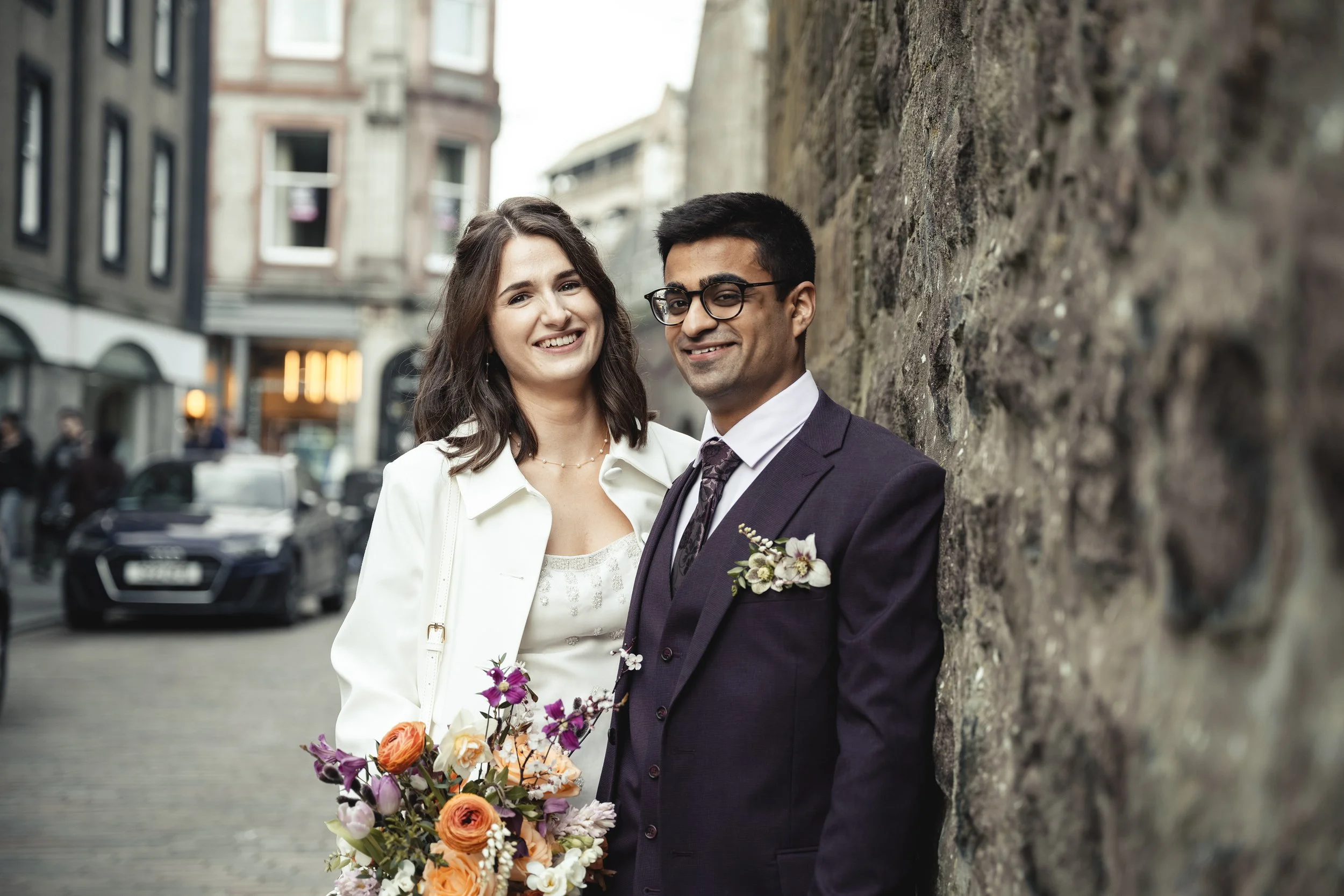A couple dressed in formal wedding attire standing outdoors near a stone wall. The woman is holding a bouquet of flowers, and the man is wearing glasses and a suit with a boutonniere. They are smiling and standing close together in an urban setting.