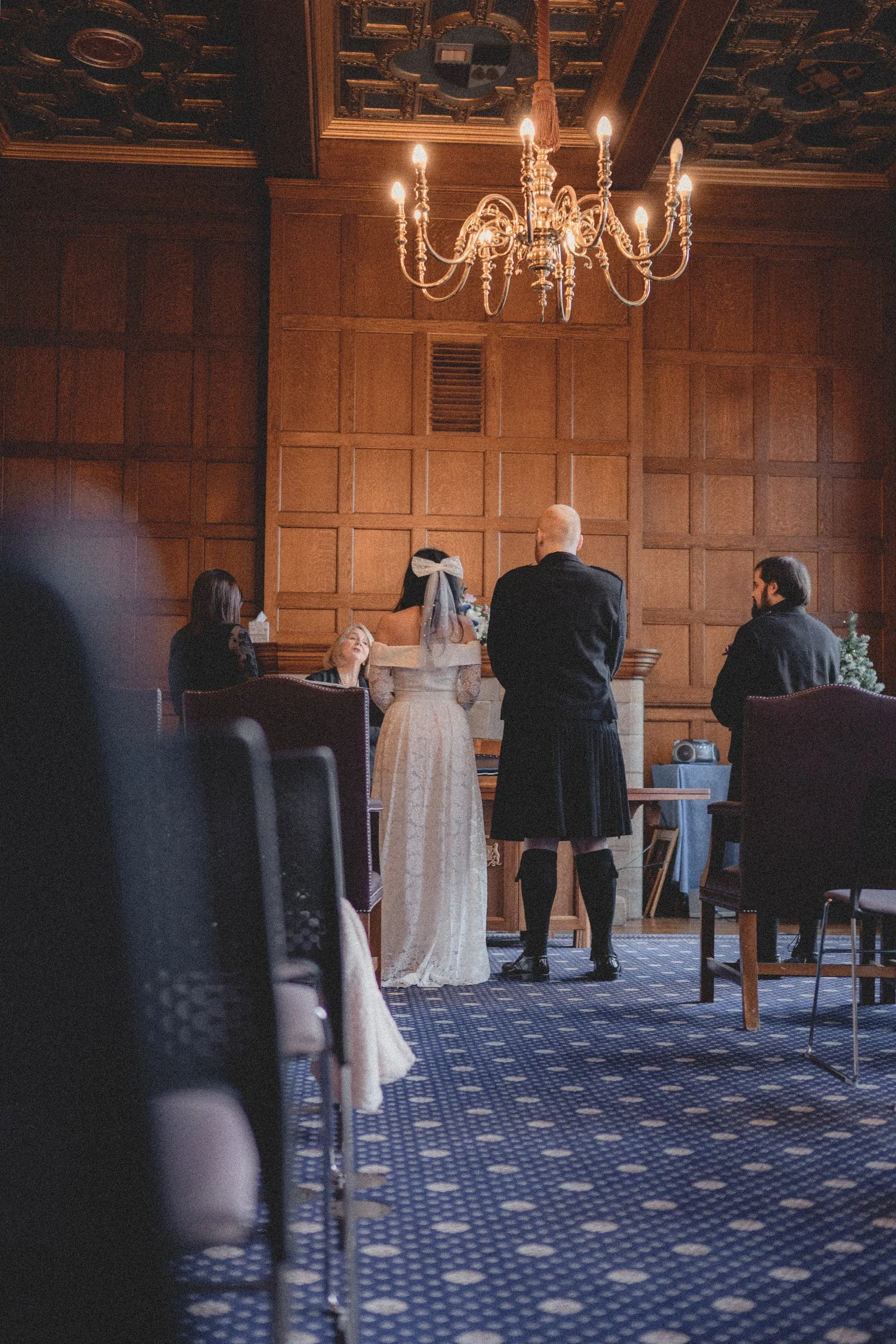 A wedding ceremony with a bride and groom standing before a judge or officiant in a wood-paneled room with a chandelier overhead.