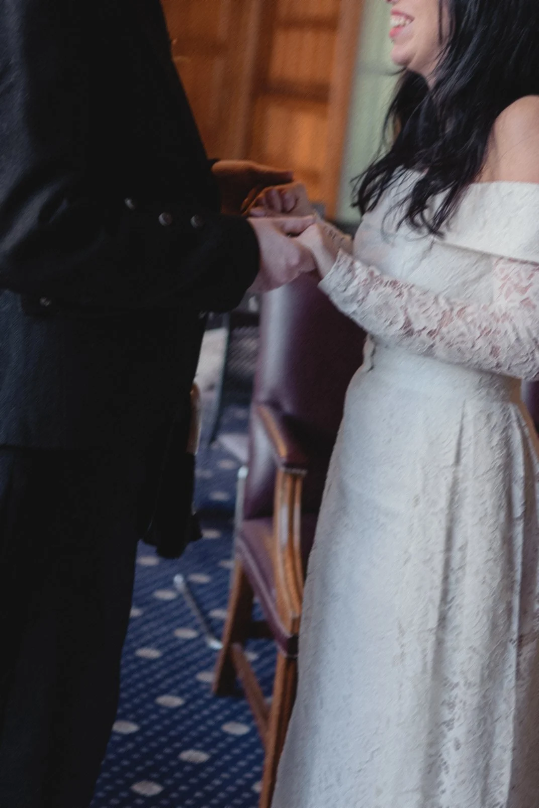 A man and woman holding hands during a wedding ceremony, with the man wearing a black suit and the woman wearing a lace wedding dress, inside a room with wooden furniture.