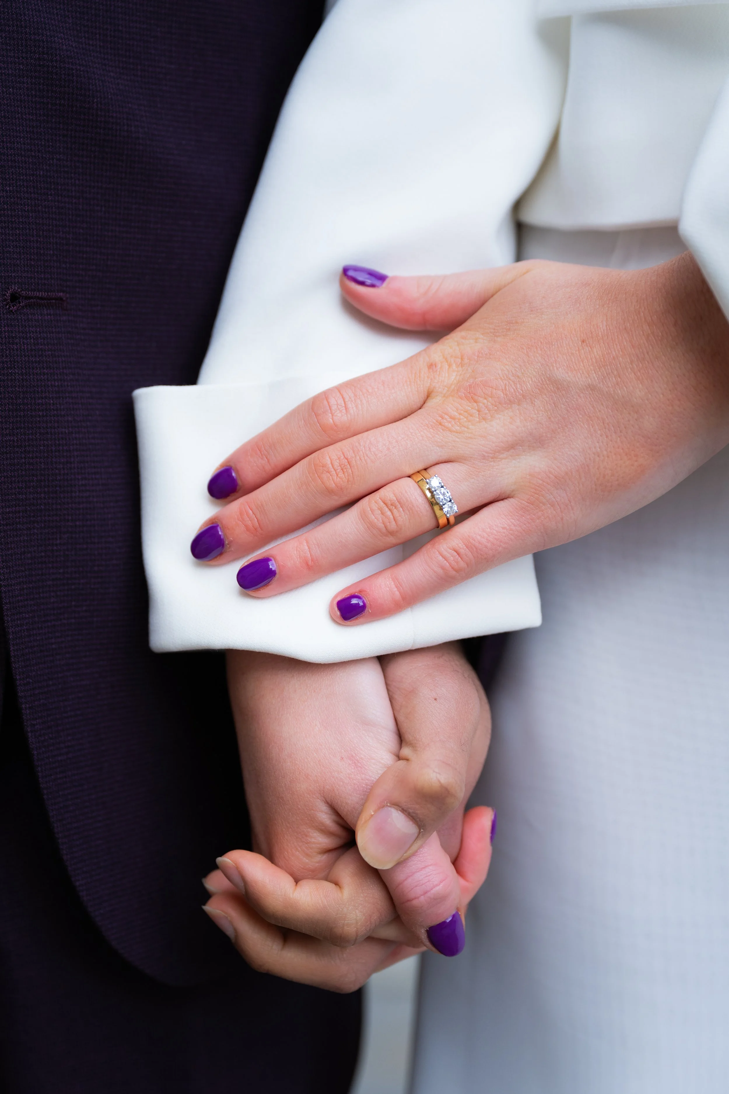 Close-up of a couple holding hands, the woman's hand resting on the man's arm, showing an engagement ring with a large diamond and purple nail polish.