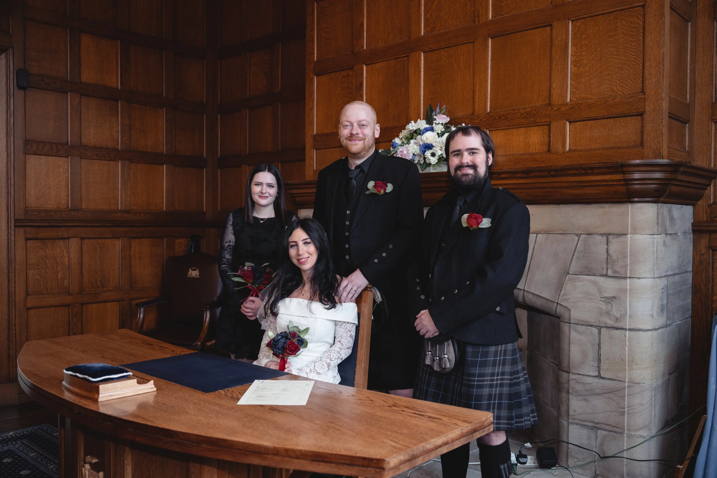 A group of five people gathered for a formal occasion in a wood-paneled room. A woman in a white dress is seated at a wooden table, holding a bouquet of red and dark blue flowers. Behind her, four others stand, including a woman in black, a tall man 
