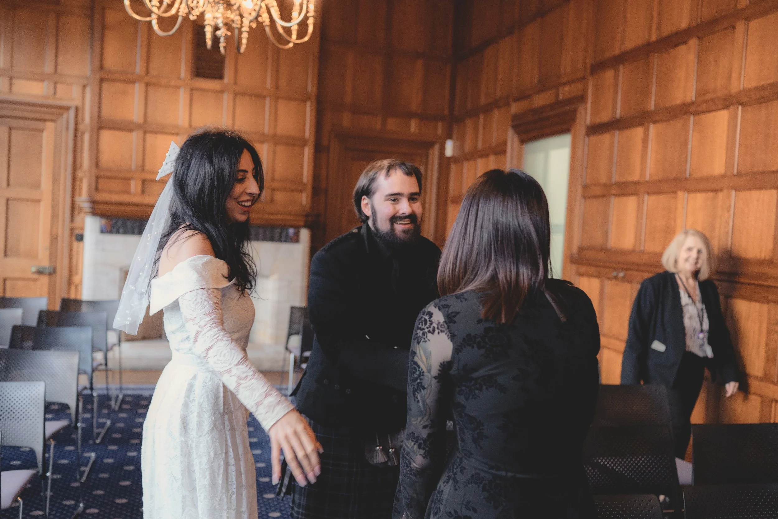 People in a wooden-paneled room engaged in conversation at a social event, with a woman in a white dress, a man with a beard, and another woman with dark hair. A woman with blonde hair is seen smiling in the background.