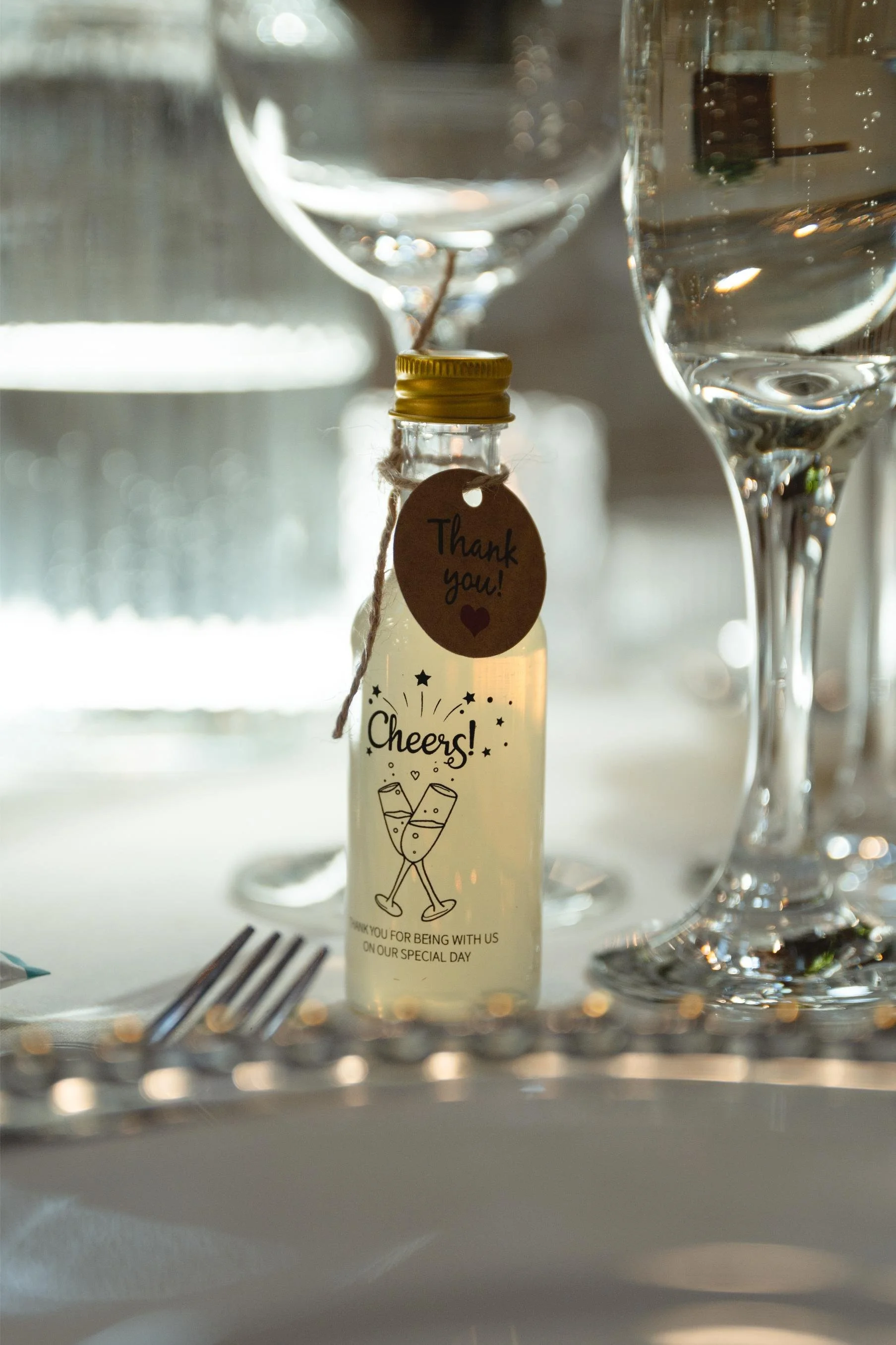 A small bottle with a yellow cap and a tag that says 'Thank you!' hanging from it, placed on a table with glassware and a white tablecloth at a celebration or special event.
