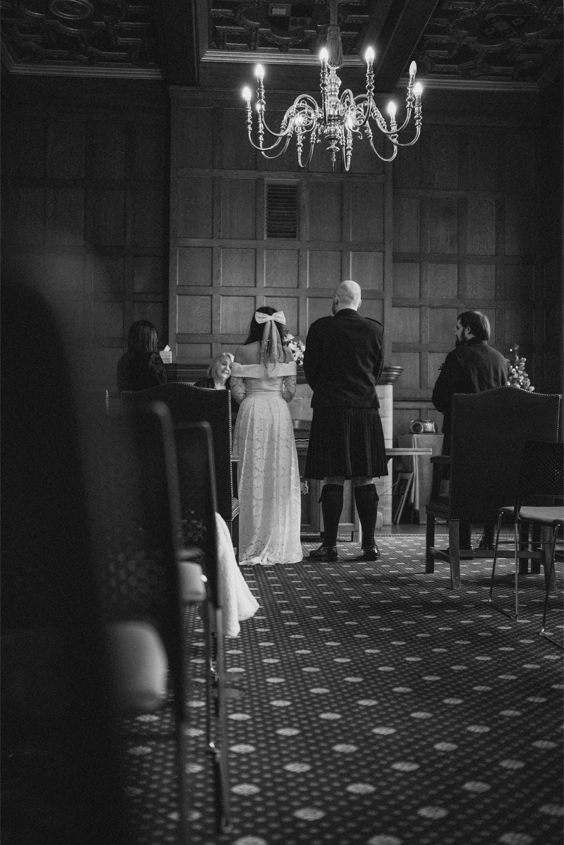 A black and white photo of a wedding ceremony inside a wood-paneled room with a chandelier. A bride in a white gown and veil stands at the altar, facing an officiant. Two men and two women stand nearby, participating in the ceremony.