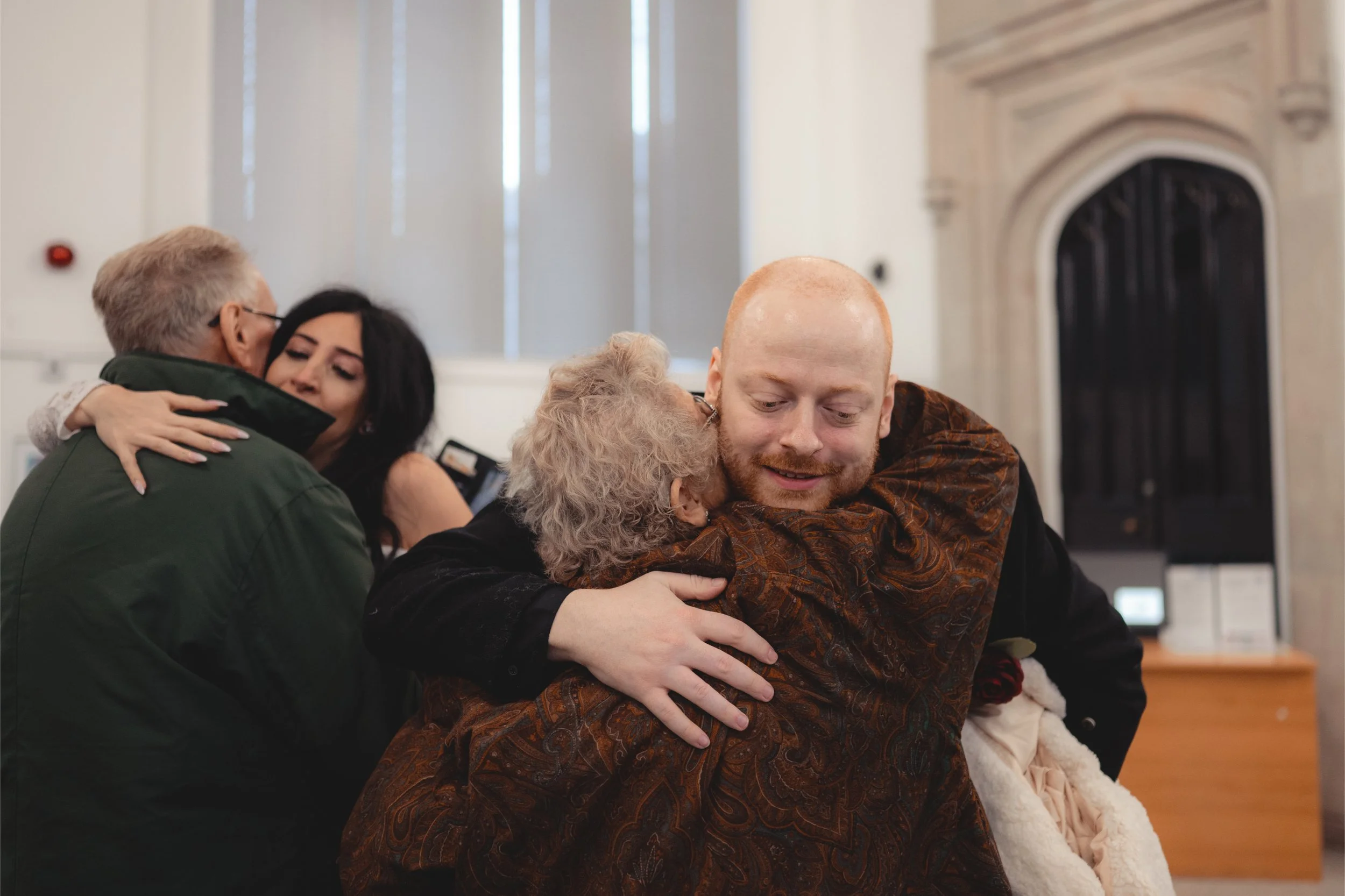 People embracing each other in a room with high ceilings, background window light, and a decorative fireplace.