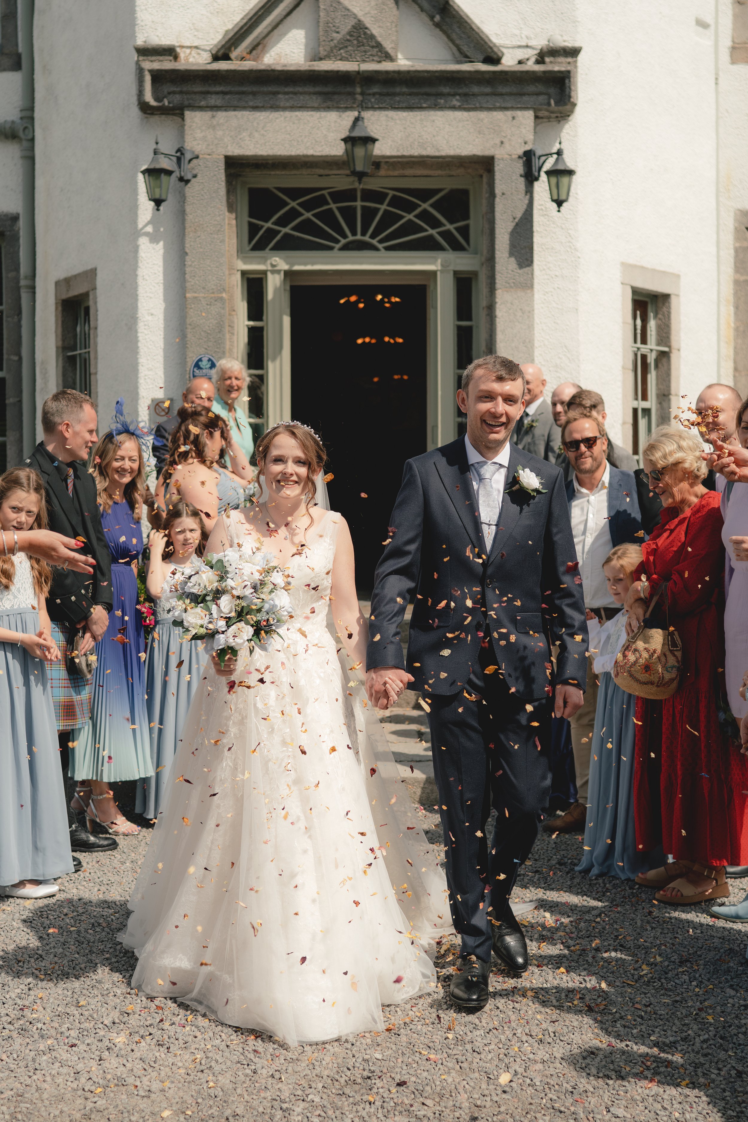 Bride and groom leaving a wedding ceremony, holding hands, surrounded by family and friends, with confetti falling around them outside a church.