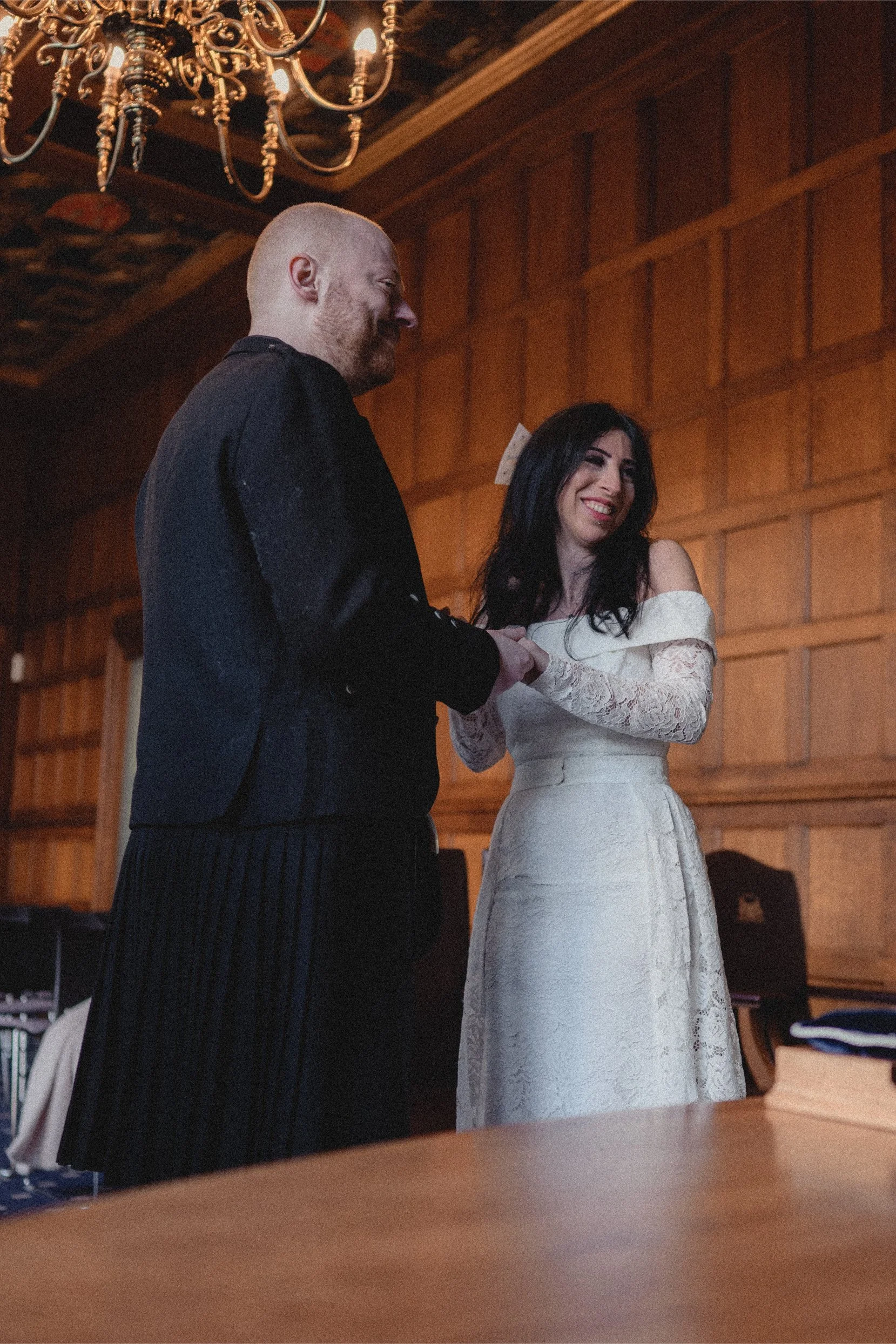 A man in a dark suit and a woman in an off-shoulder lace wedding dress holding hands and smiling at each other in a wood-paneled room with a chandelier overhead.
