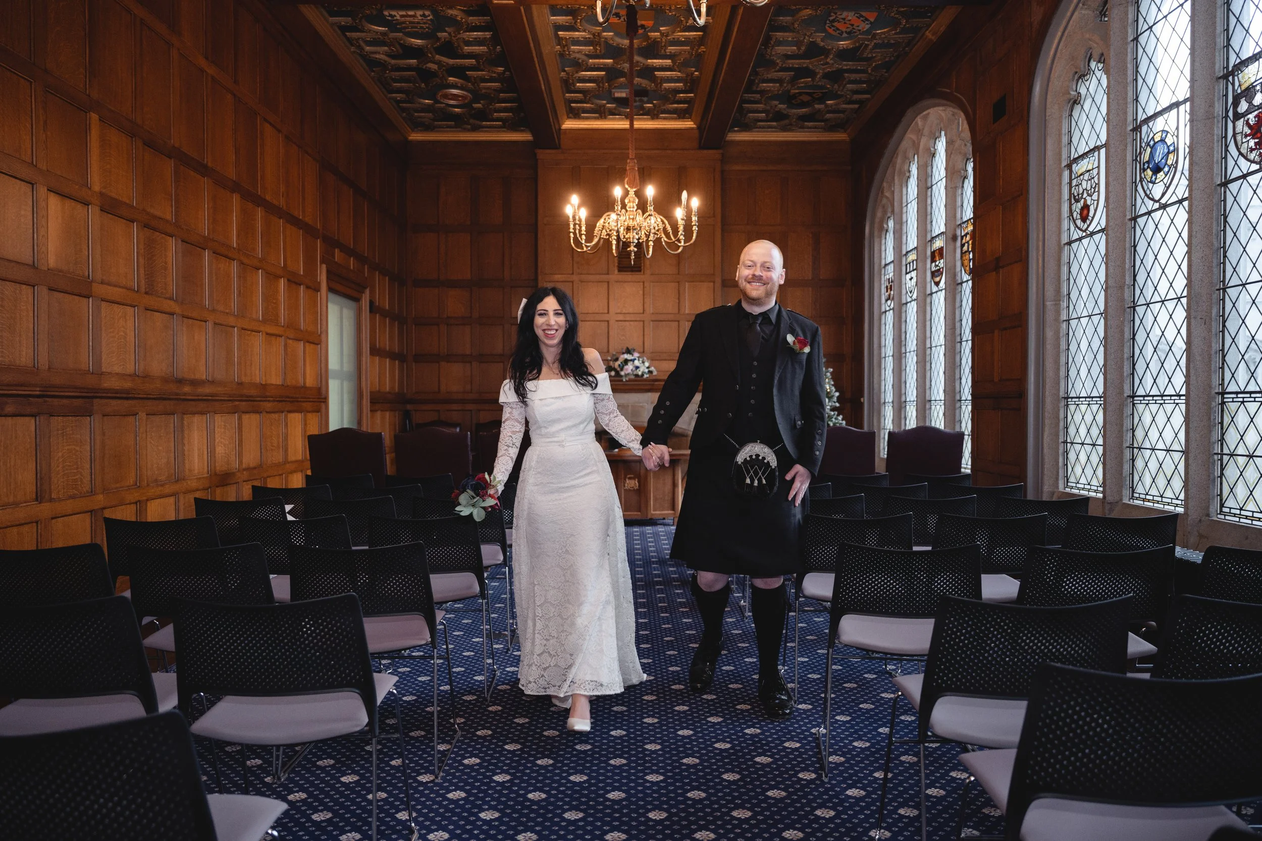 A bride and groom walking hand in hand down the aisle in a wooden-paneled wedding ceremony room with stained glass windows, chandelier, and empty chairs.