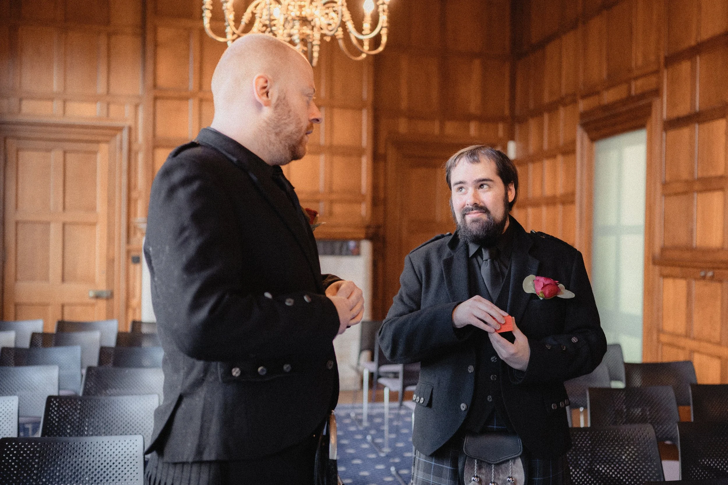 Two men in formal attire, one with a boutonnière and kilt, standing and talking inside a wood-paneled room with a chandelier, chairs, and a doorway.