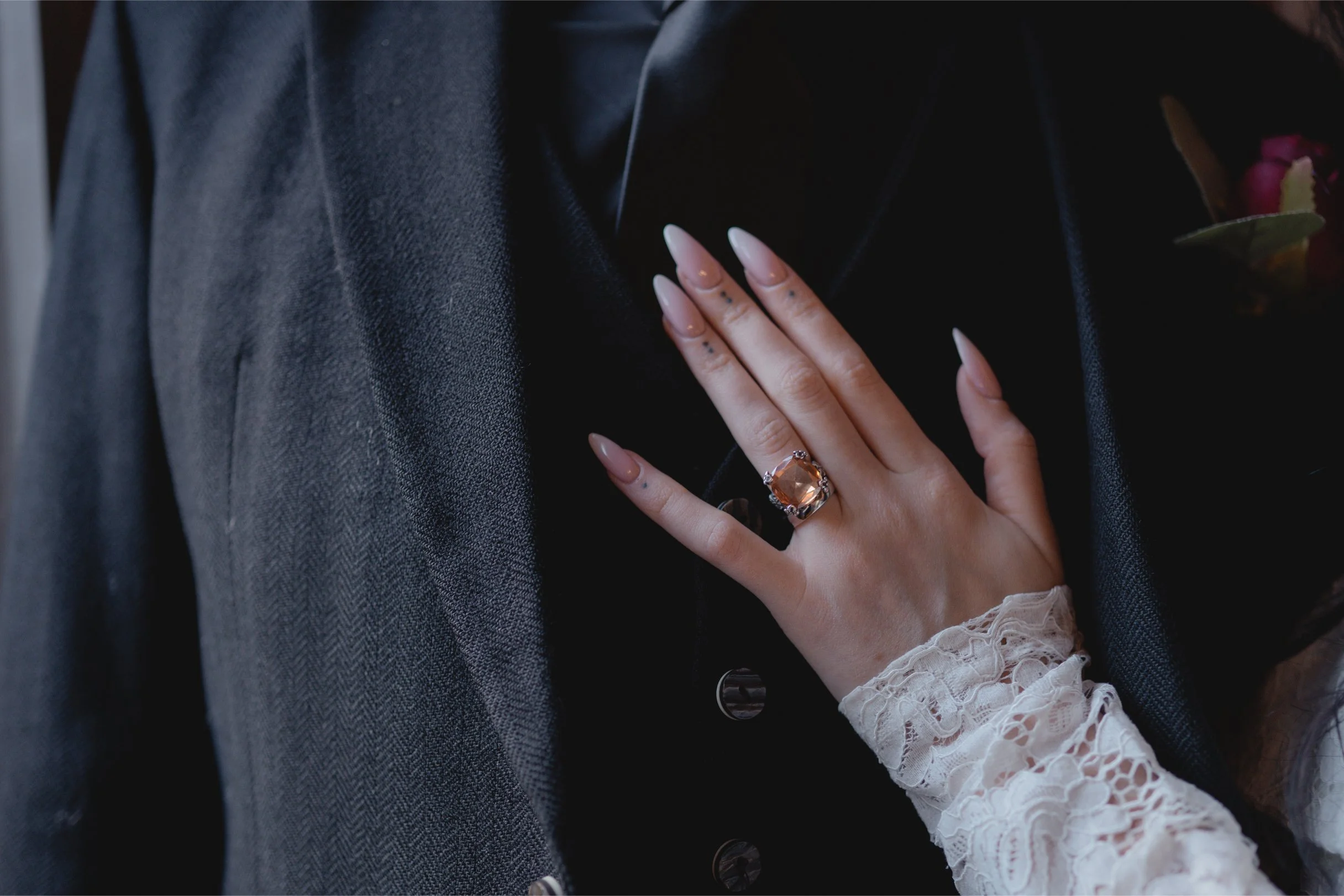 A woman's hand resting on a man's chest, wearing a large gemstone ring and a lace sleeve.