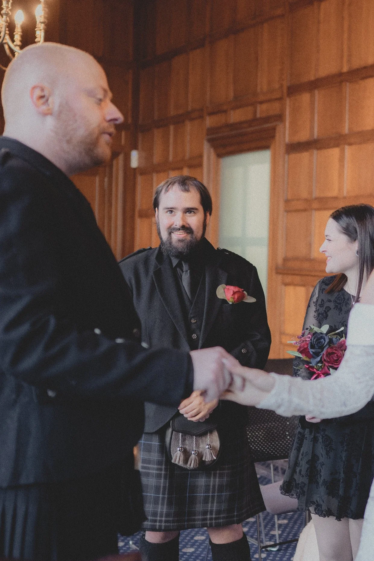 A wedding ceremony with a groom in a kilt shaking hands with a guest, while a man and woman look on smiling in a wood-paneled room.