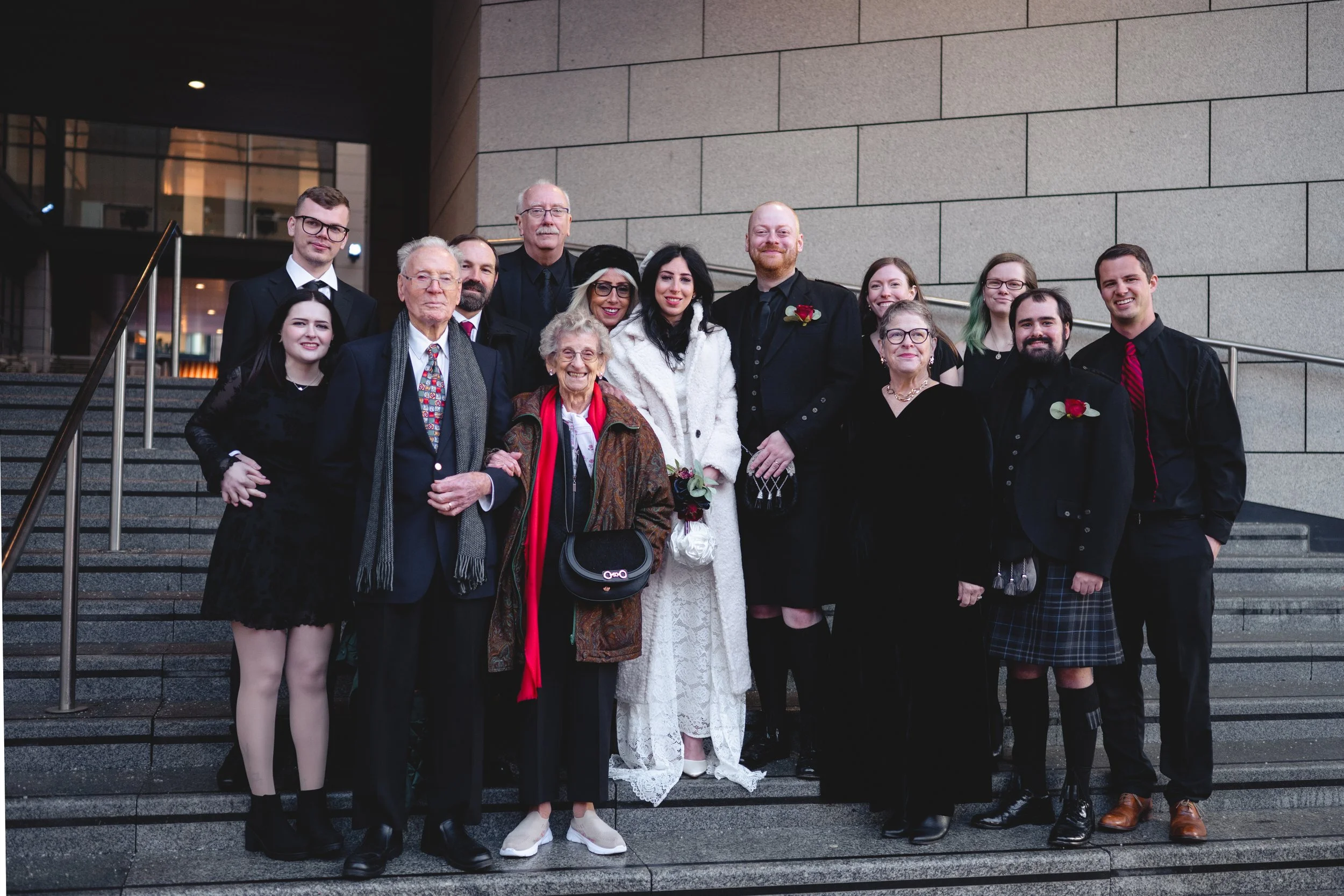 A large group of people, including a bride in a white dress and groom in a black suit with a red rose boutonniere, poses on the steps outside a modern building during a wedding celebration.
