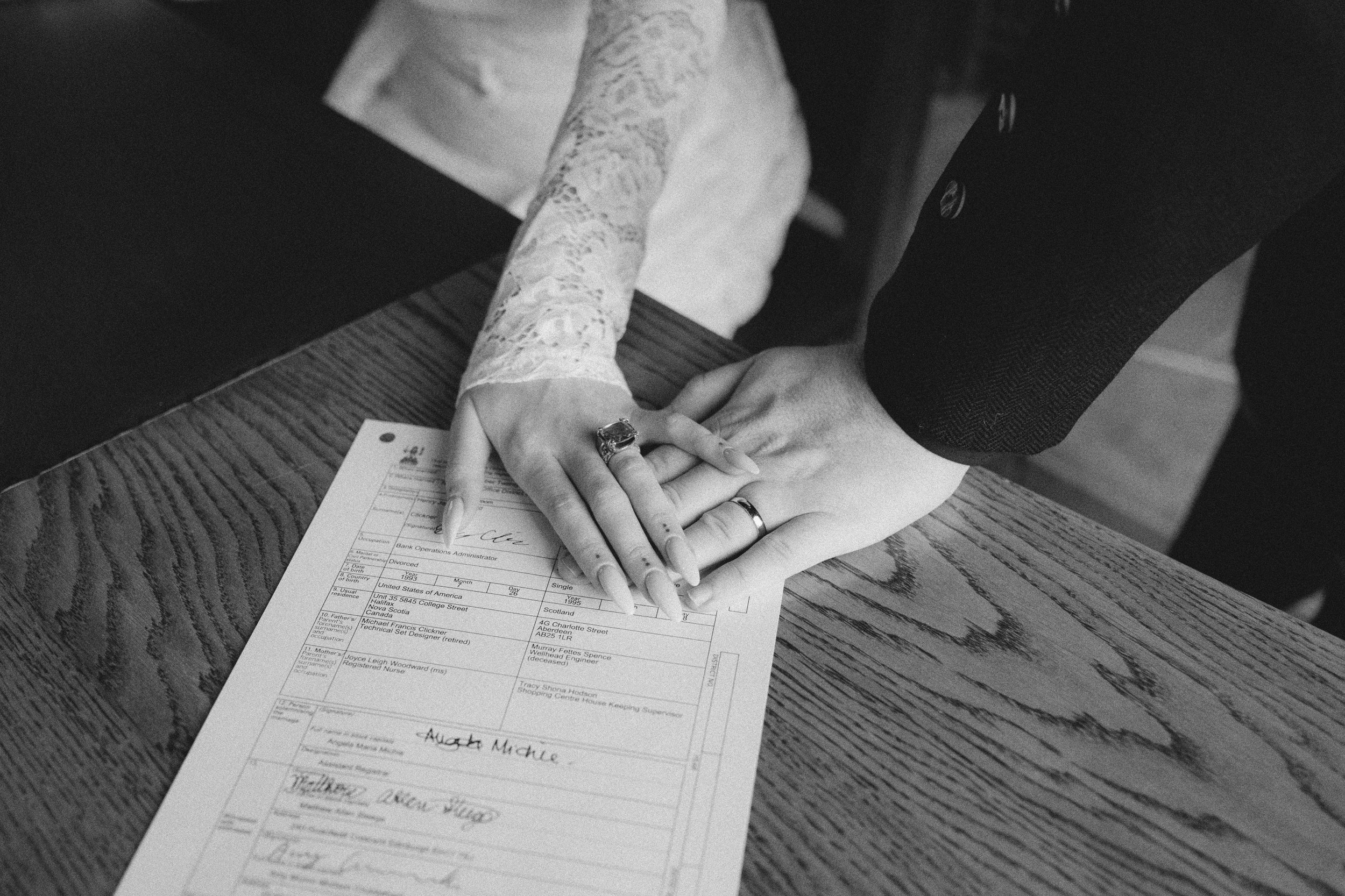 Hands of a woman and a man are clasped over a document on a wooden table, symbolizing a wedding or legal agreement, with the woman's hand showing an engagement ring and the man wearing a wedding band.