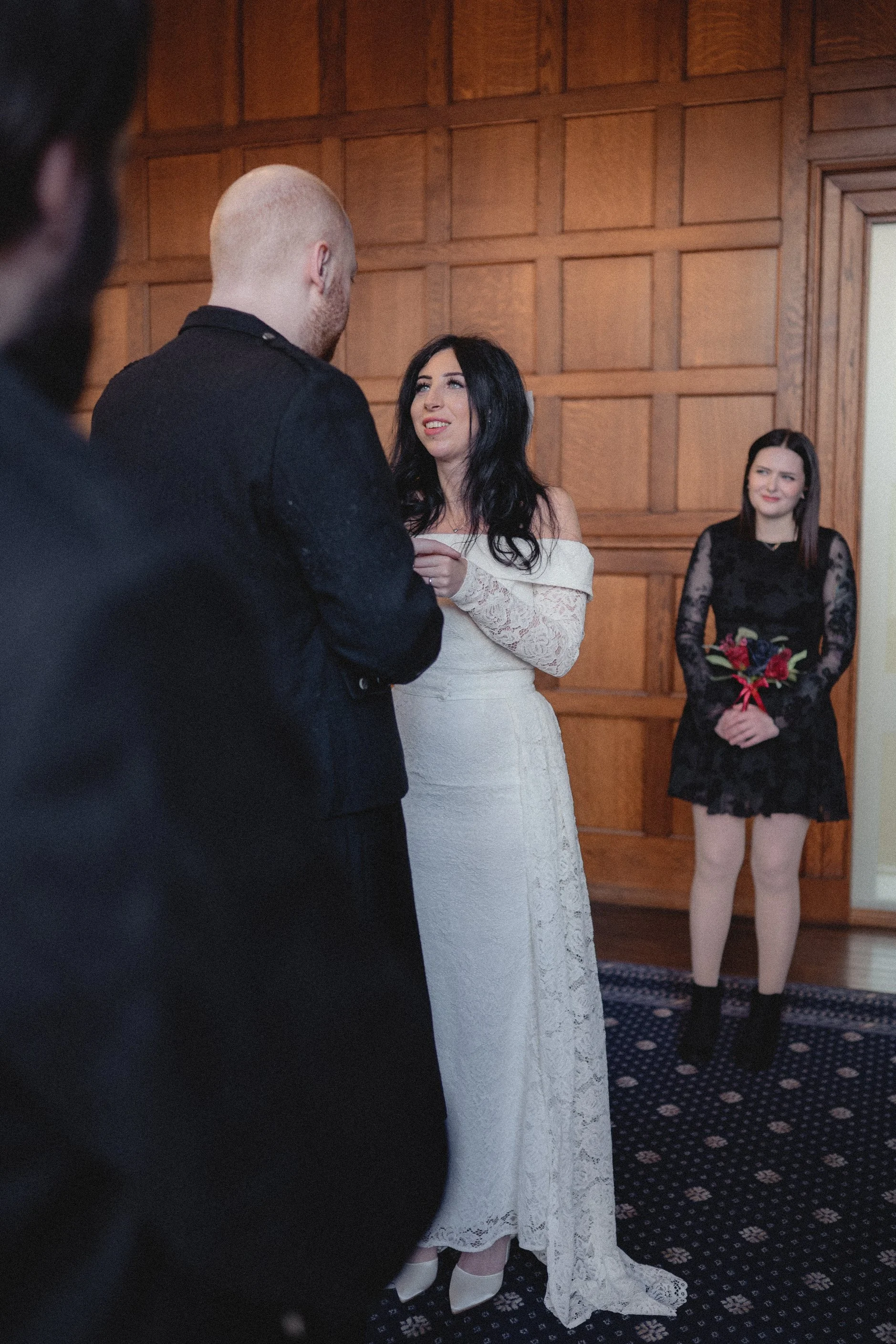 A woman in an off-the-shoulder lace wedding dress is having a moment with a man during a wedding ceremony, with a bridesmaid holding a bouquet in the background.
