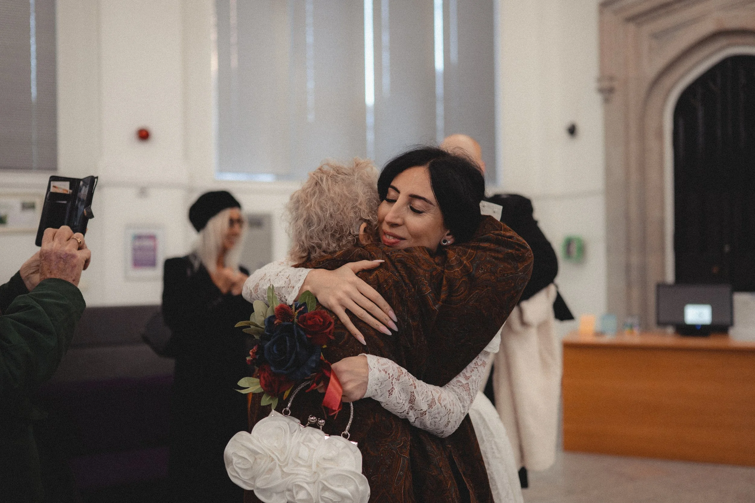 Two women hugging each other warmly at a social gathering, with one holding a bouquet of flowers and a white bag. There are other guests in the background, and a person on the left is taking a photo of the embrace.