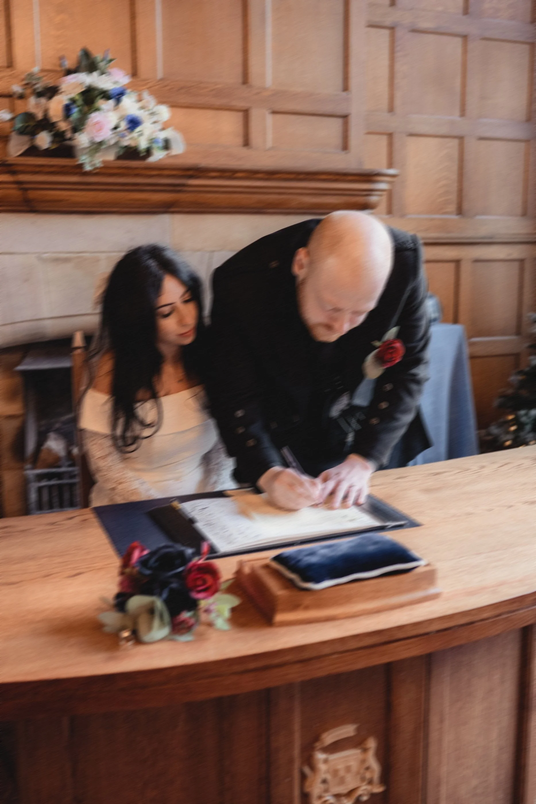 A woman in a white dress sits at a wooden table while an older man signs a document for her. There are flowers and a pillow on the table, with a wood-paneled wall in the background.