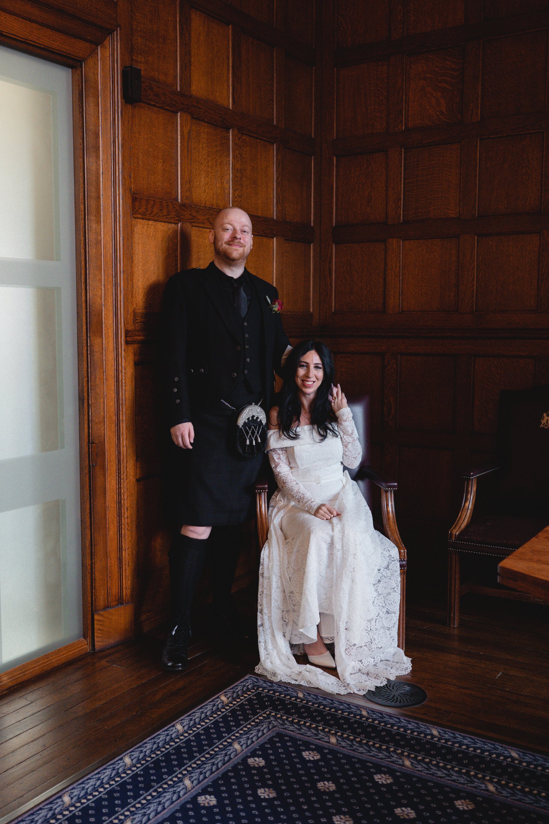 A man in traditional Scottish attire and a woman in a wedding dress pose for a photo in a room with wooden walls. The woman is seated and smiling, while the man stands beside her, smiling slightly.