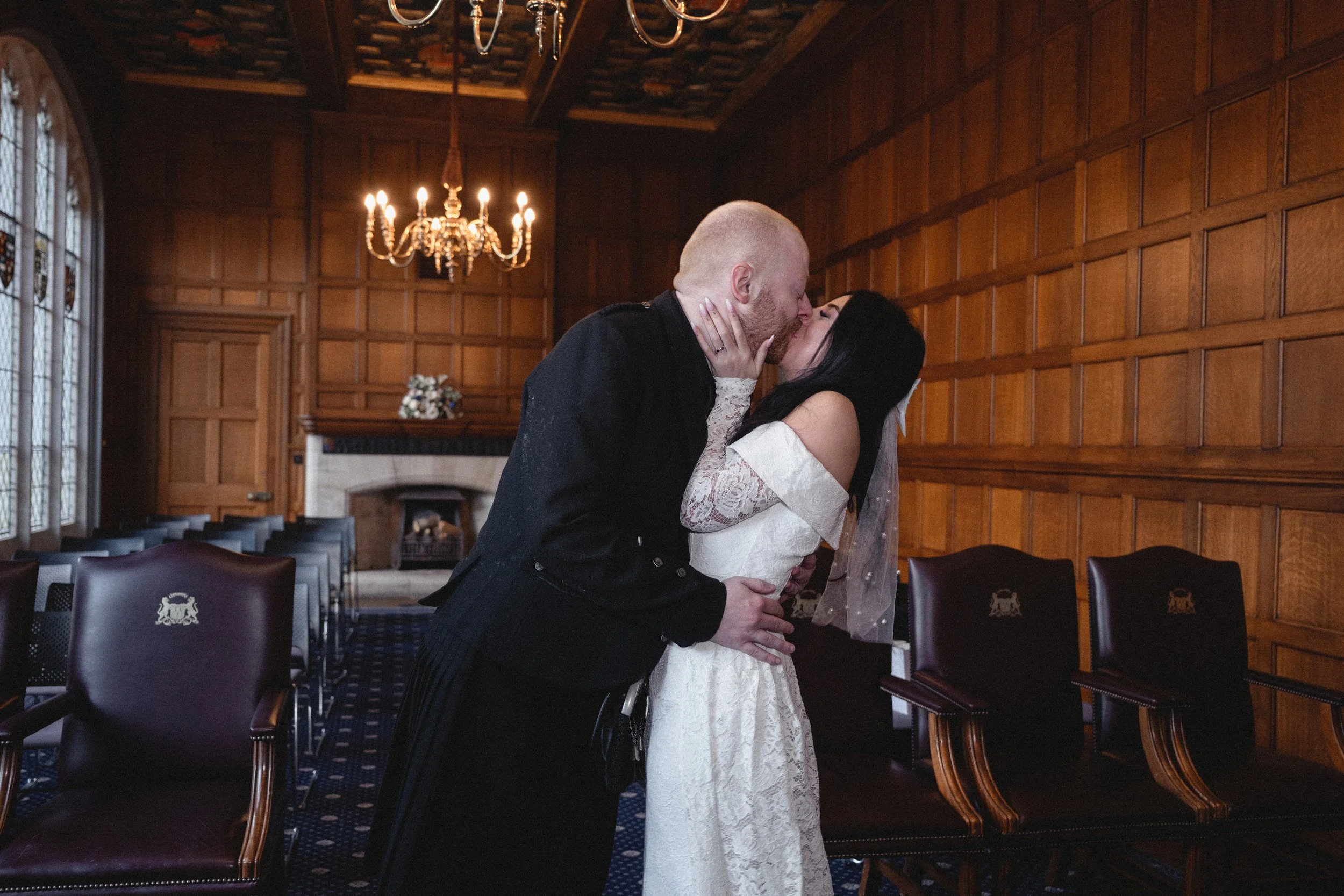 A couple shares a kiss in a wood-paneled room with stained glass windows and a chandelier.