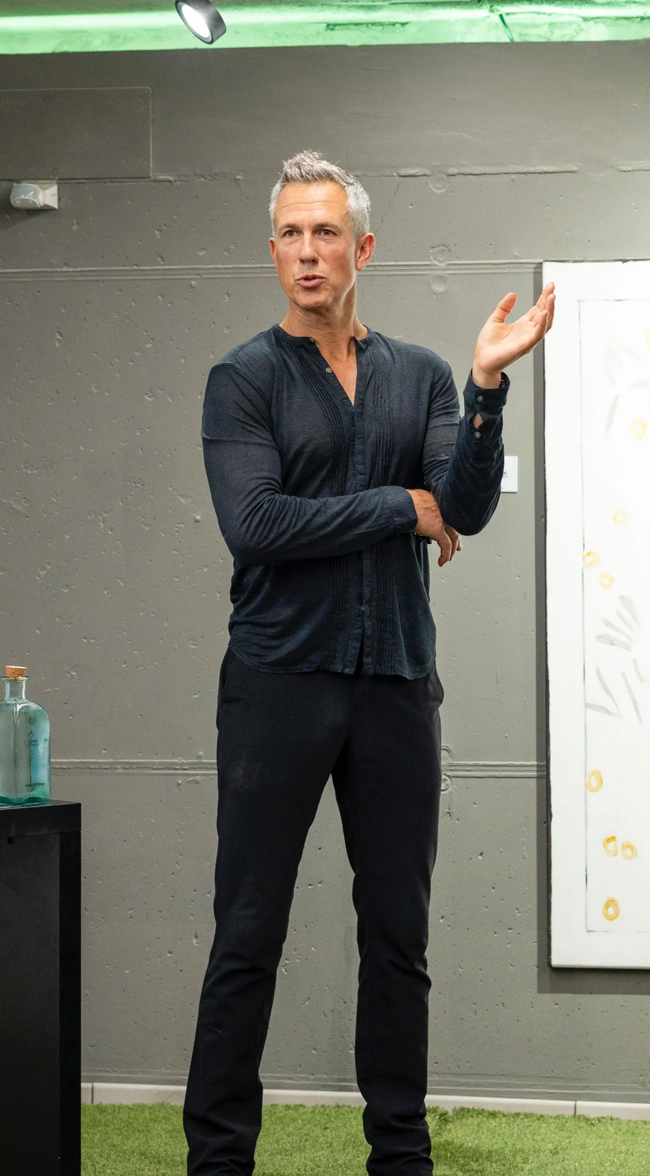 A man with gray hair in a black long-sleeve shirt and black pants standing in front of a gray wall, gesturing with his right hand, possibly giving a presentation.