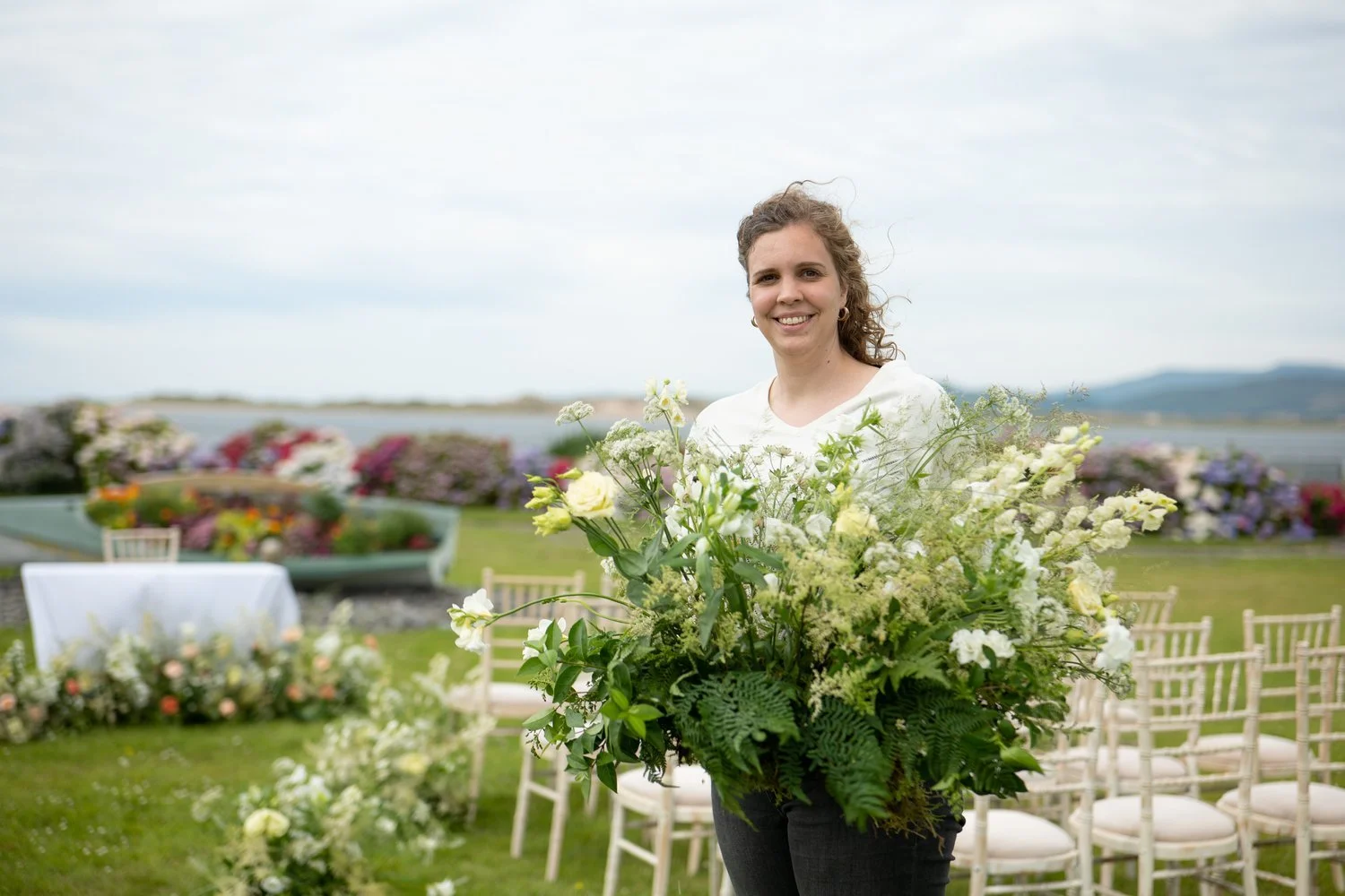 Jiska Healy holding a large floral arrangement at an outdoor event setup with chairs and scenery in the background.