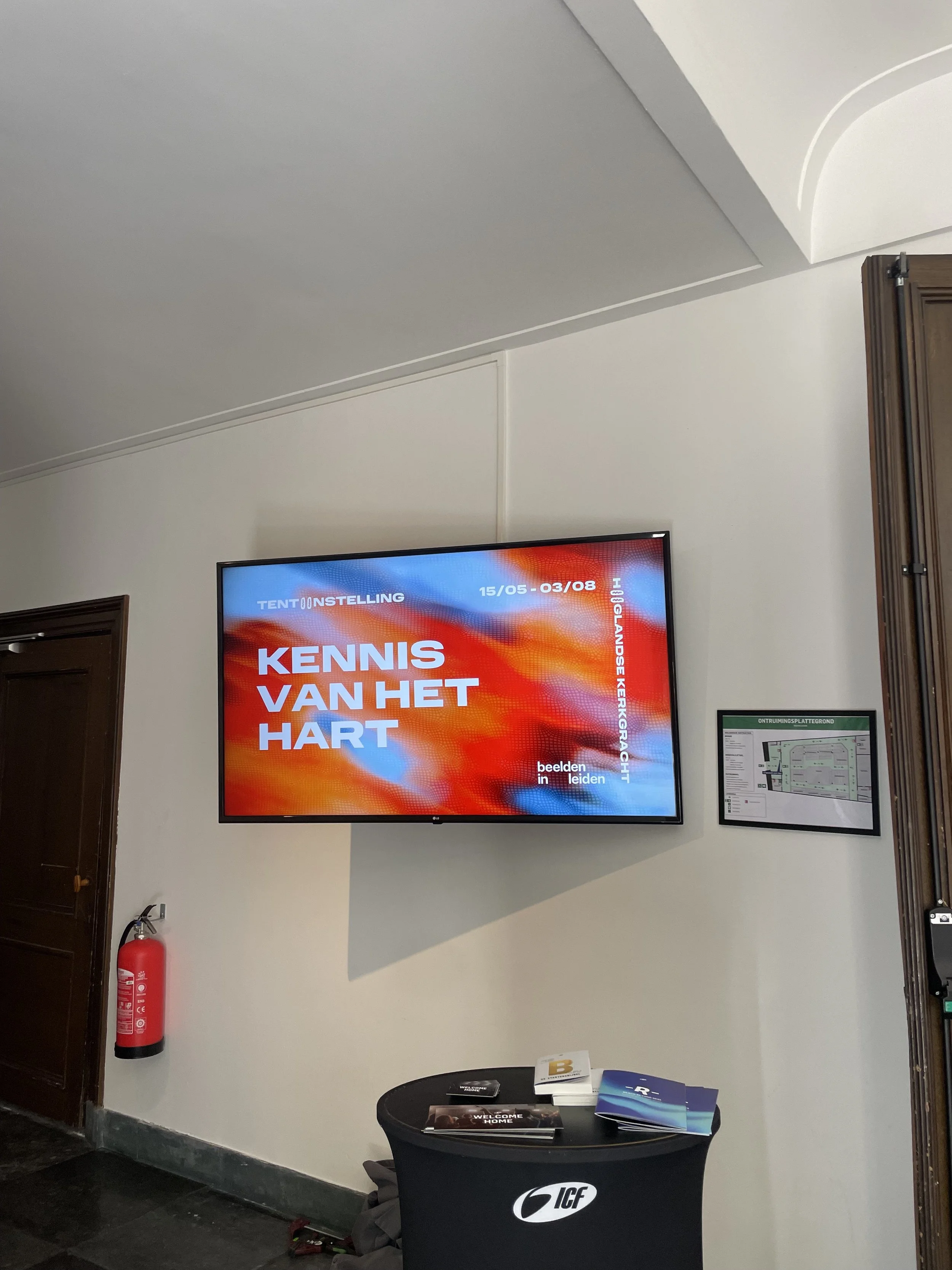 Een digitale scherm met de tekst "KENNIS VAN HET HART" en een datum van 15/05 - 03/08, hangt aan een witte muur. Naast het scherm bevindt zich een klein bestuursbord, onder een ronde tafel met brochures en een zwart object, een brandblusapparaat aan