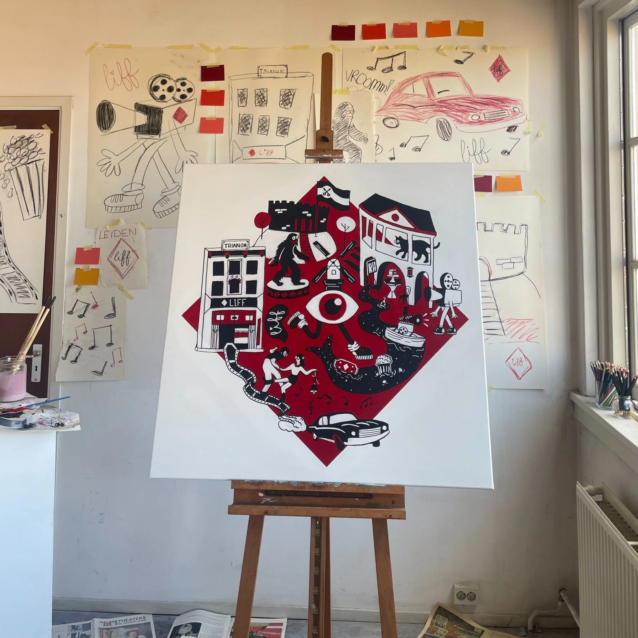 Een kunstwerk op een ezel met een zwart, rood en wit ontwerp van een stad met gebouwen, mensen, dieren, auto’s en muziekelementen.