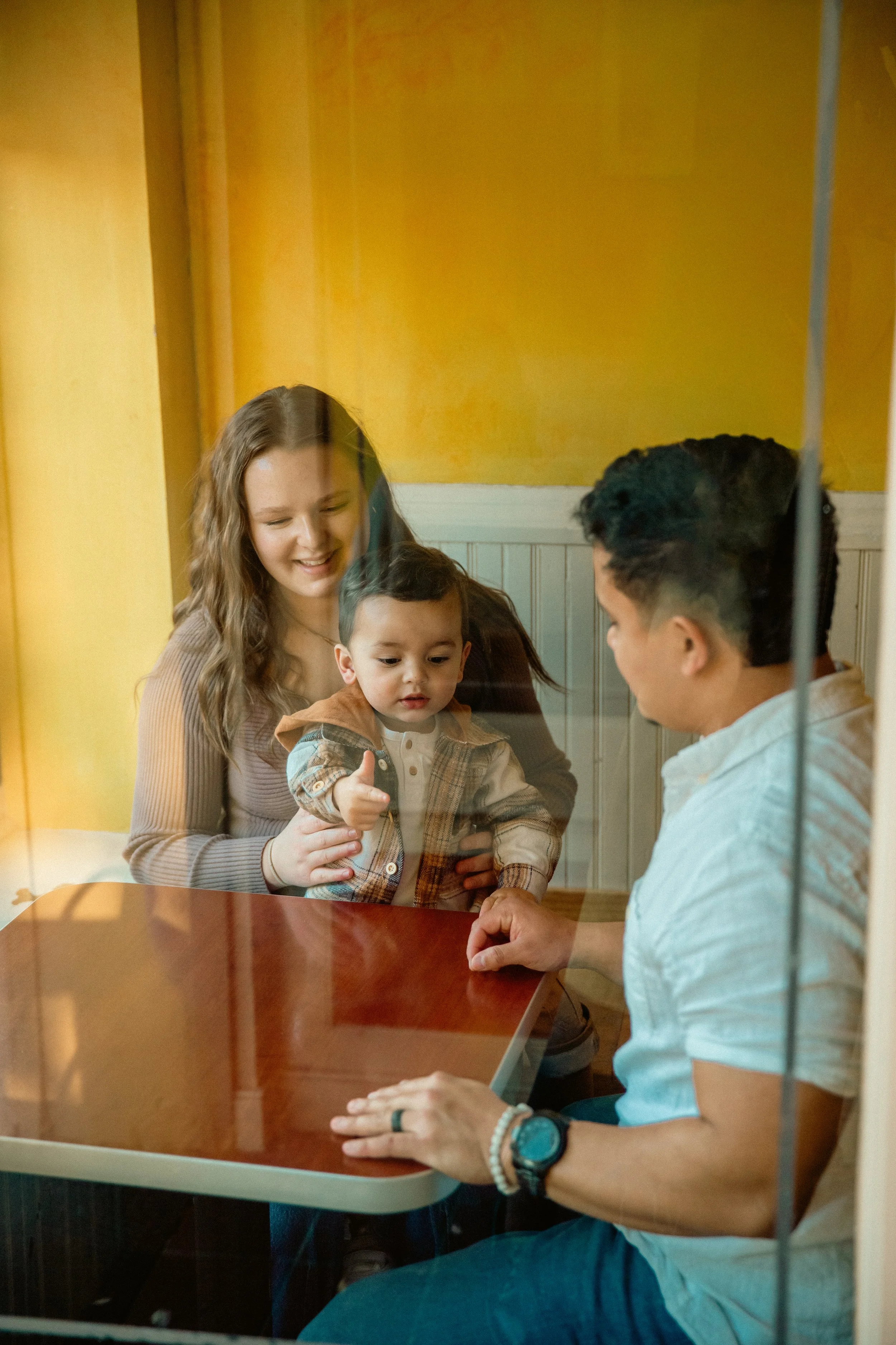 A family with a woman, a young child giving a thumbs-up, and a man sitting at a table, seen through a glass window.