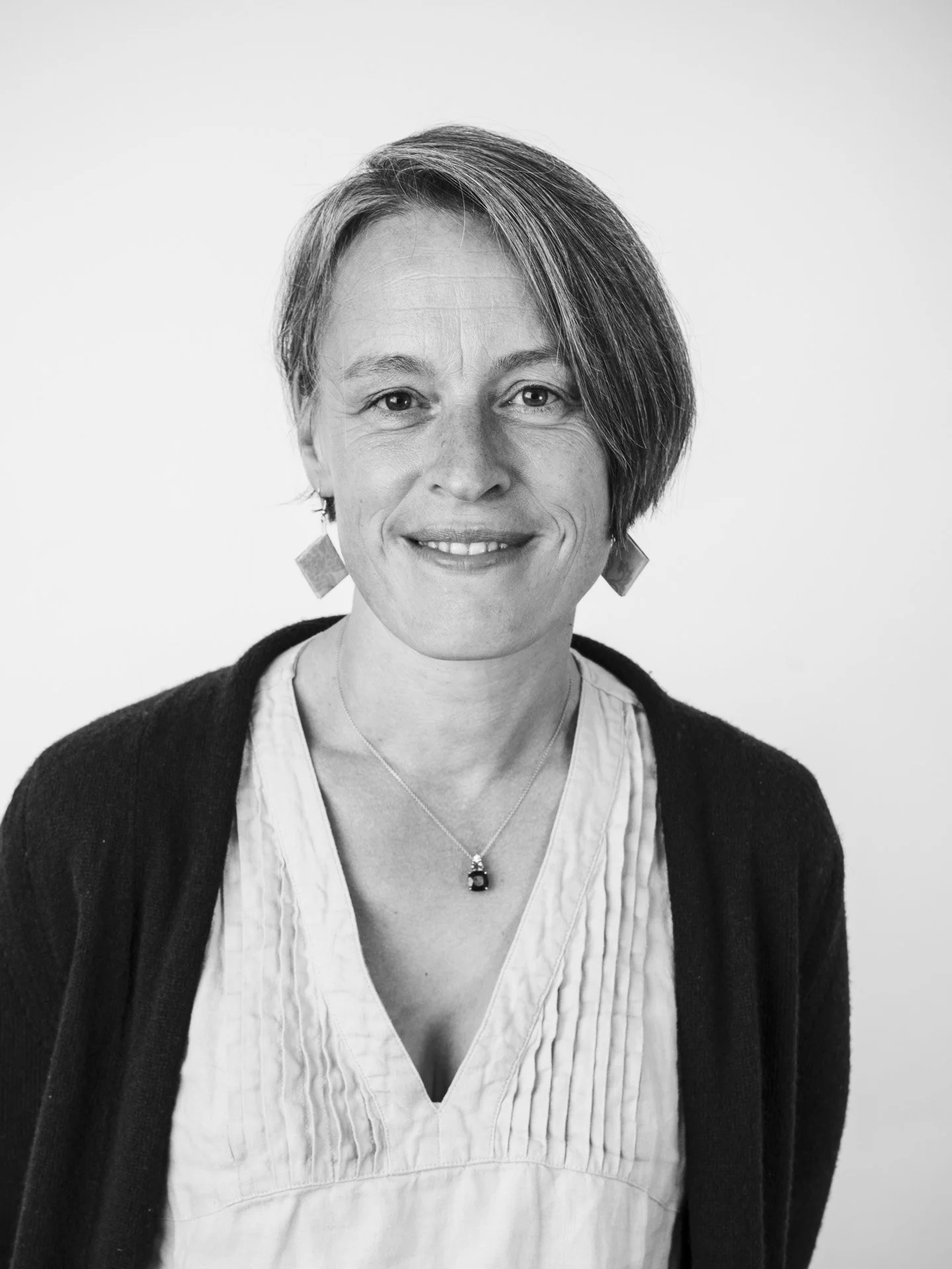 Portrait en noir et blanc d'une femme souriante portant un col en V, un cardigan noir, et des boucles d'oreilles géométriques, avec un collier orné d'une pierre