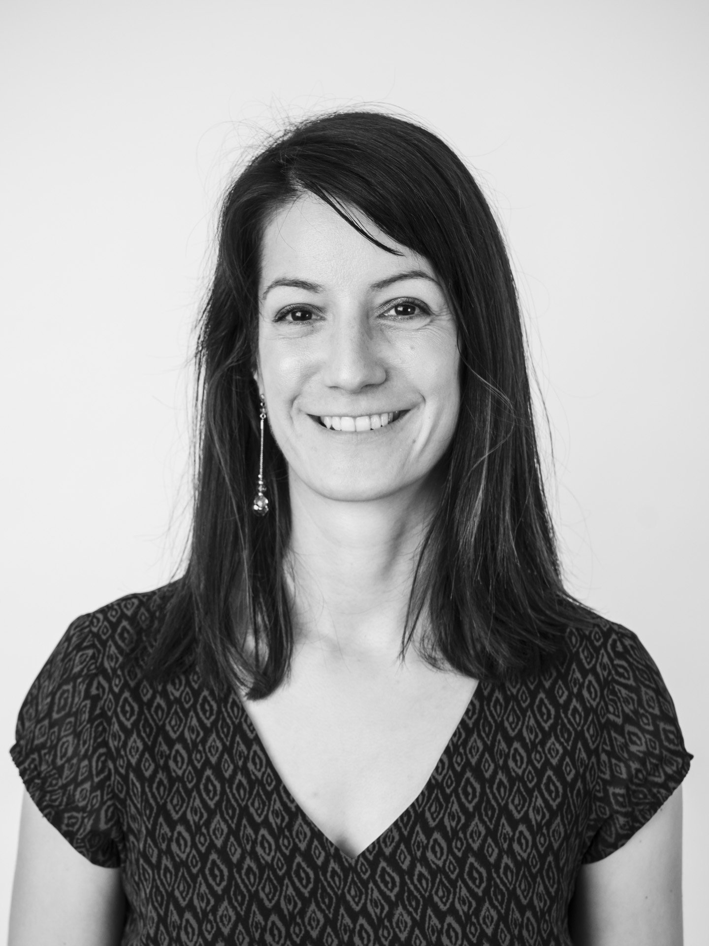 Portrait en noir et blanc d'une femme souriante avec des cheveux bruns et longs, portant une robe à motifs géométriques et une boucle d'oreille pendante.