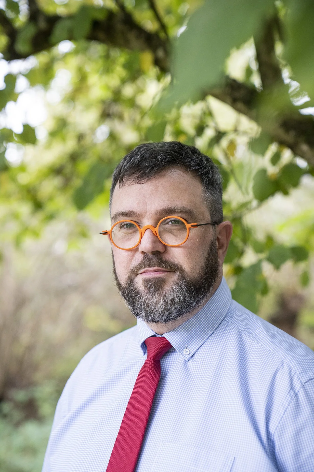 Un homme avec une barbe, portant des lunettes orange, une chemise à carreaux bleue et une cravate rouge, pose à l'extérieur devant des feuilles vertes.