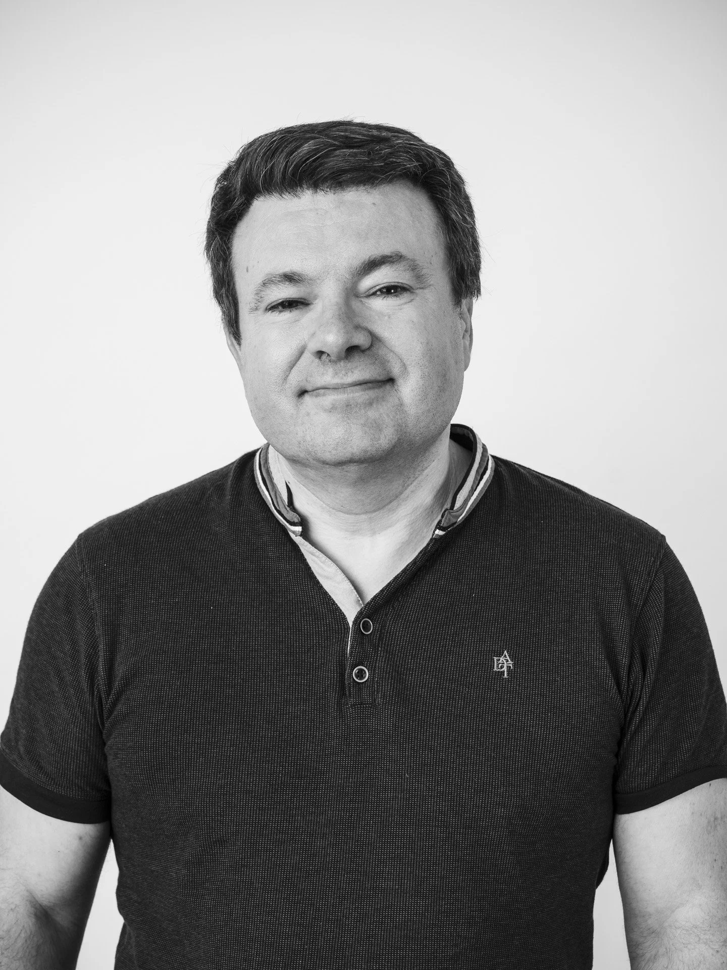 Un portrait en noir et blanc d'un homme souriant portant un polo à col ras du cou, contre un fond blanc.