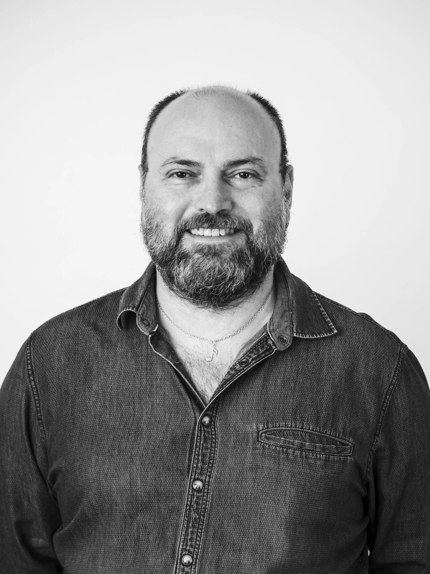 Portrait en noir et blanc d'un homme avec barbe et cheveux ras, portant une chemise en denim et une chaîne