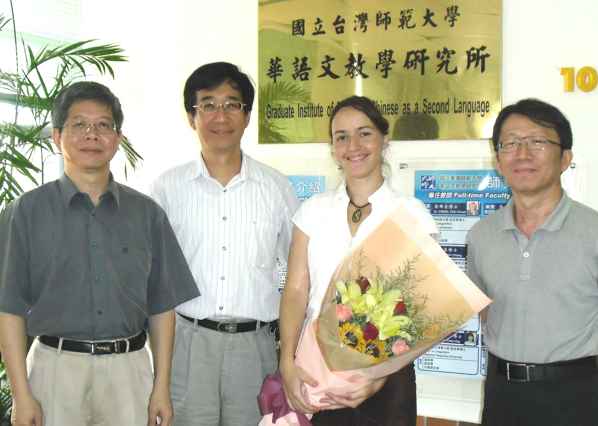 Four people standing in front of a sign that reads 'Graduate Institute of Chinese as a Second Language' at a university in Taiwan. One person is holding a bouquet of flowers.