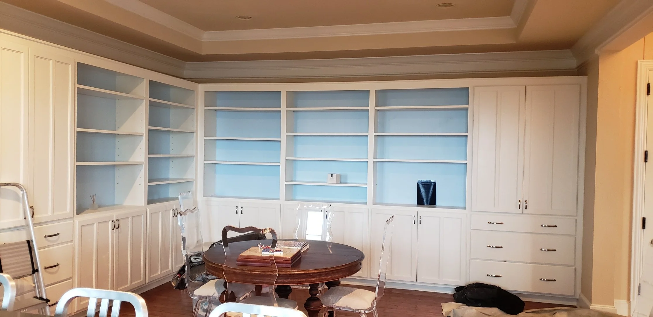 Empty built-in white cabinet with open shelves and closed drawers in a room, with a round wooden table and transparent chairs in front, and a ladder and black bag on the floor.