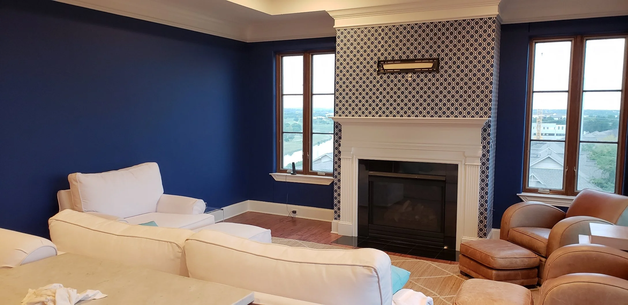 A living room with blue walls, a fireplace with a patterned wallpaper, large windows, a white sofa, a tan armchair with an ottoman, and a view of neighboring houses and a river outside.