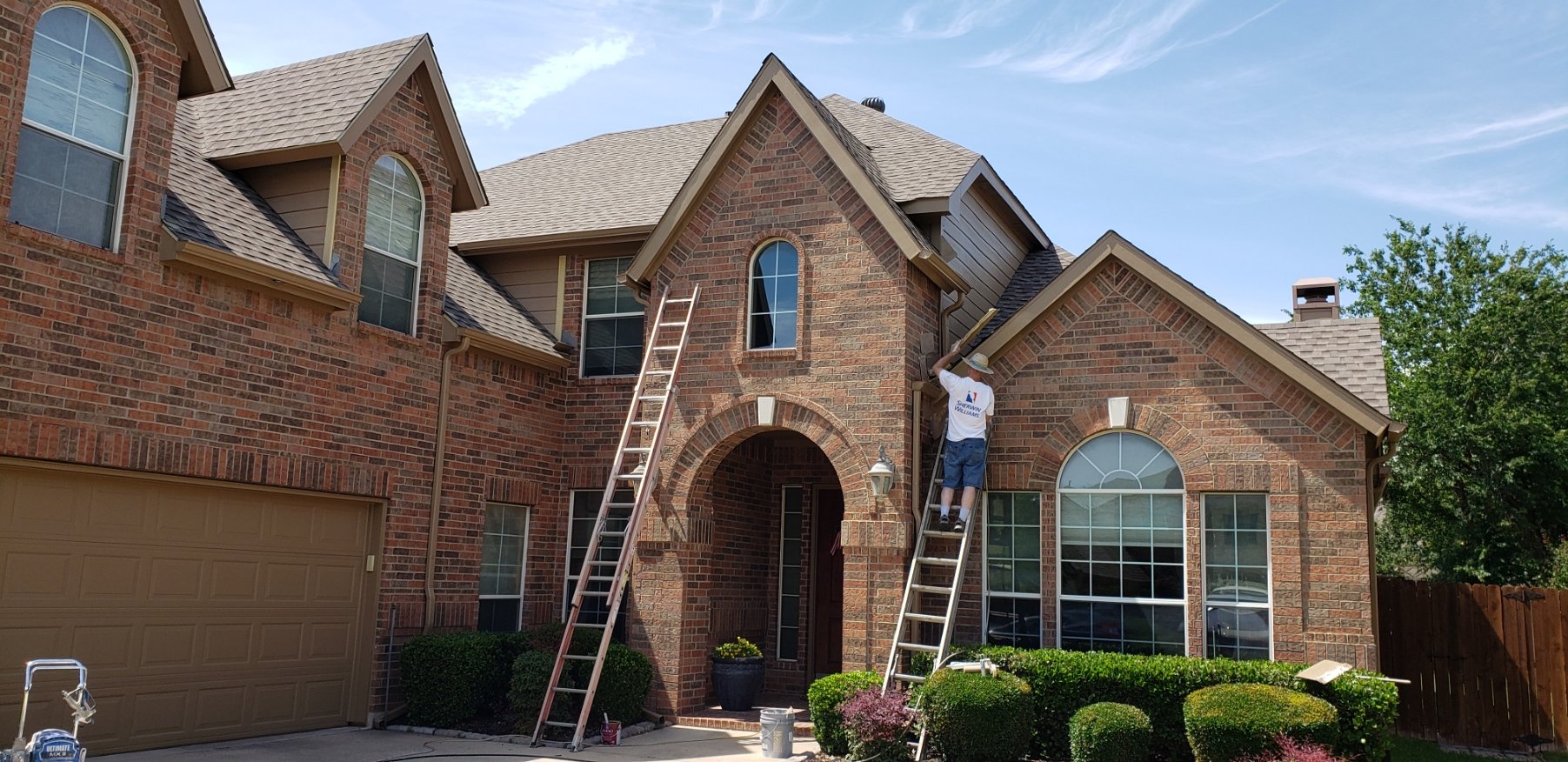 Two workers on ladders working on the roof and gutter of a large brick house during daytime. The house has multiple peaked roofs, large arched windows, and a tan garage door. There are bushes and plants in front of the house.